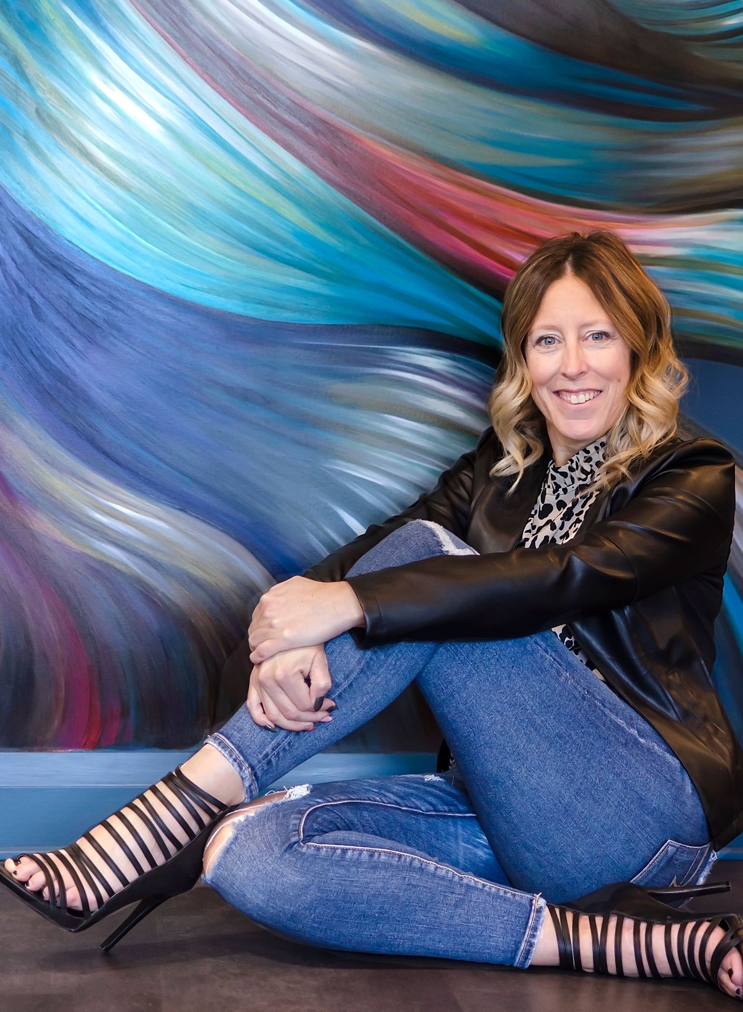 Smiling woman in leather jacket and jeans sits against vibrant colorful swirl art backdrop.