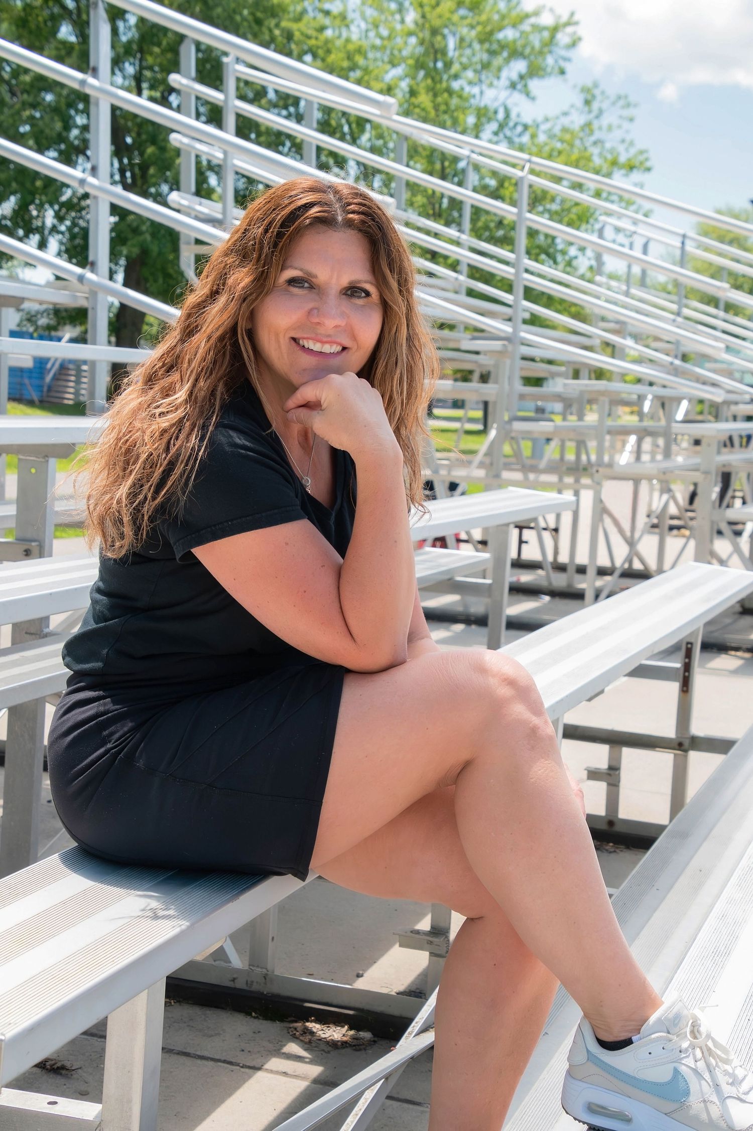 Smiling woman with long red hair sitting on bleachers in black dress outdoors.