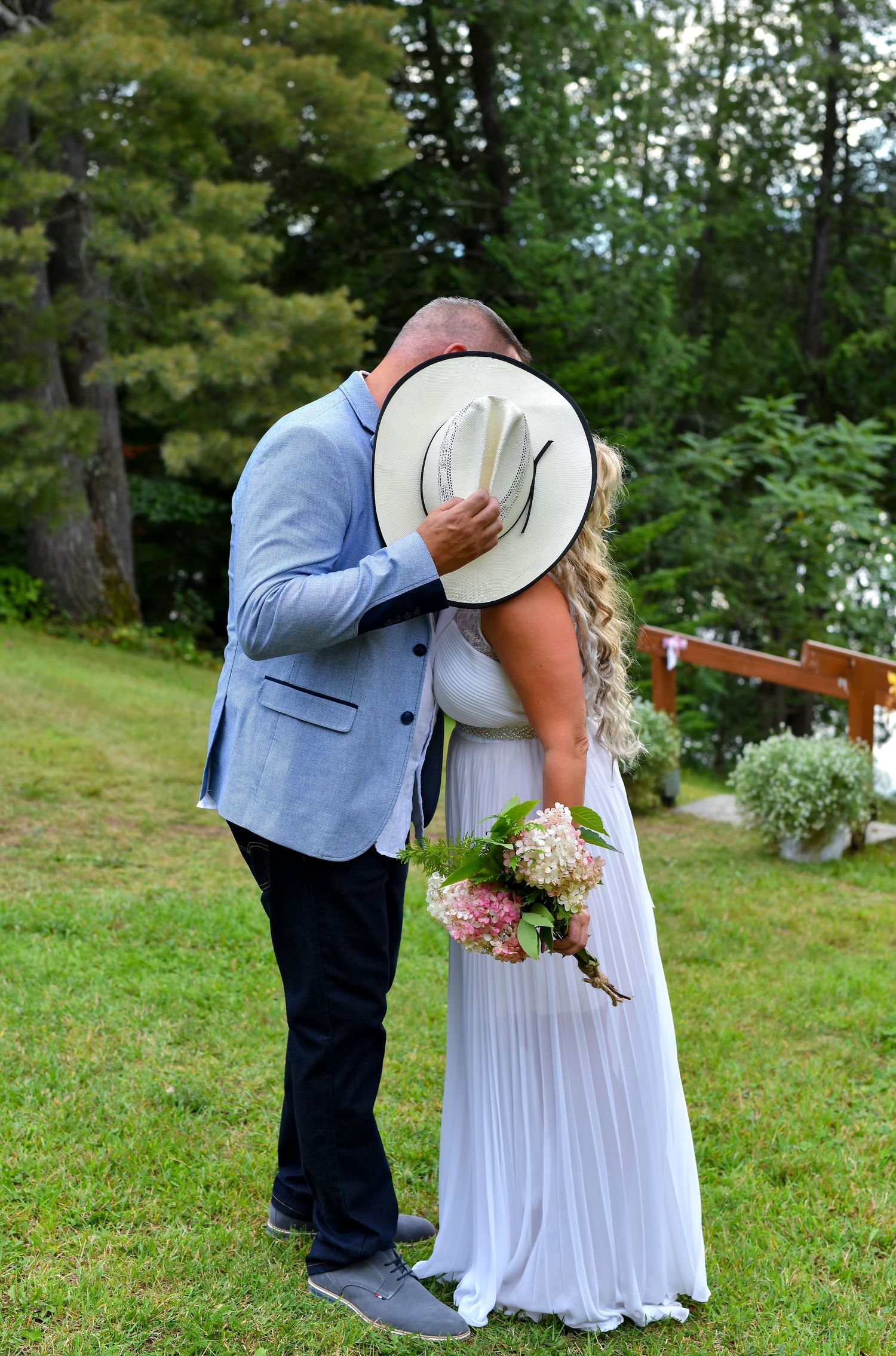 Newlyweds sharing a first kiss hidden behind a white hat, bride holding pink hydrangea bouquet outdoors.