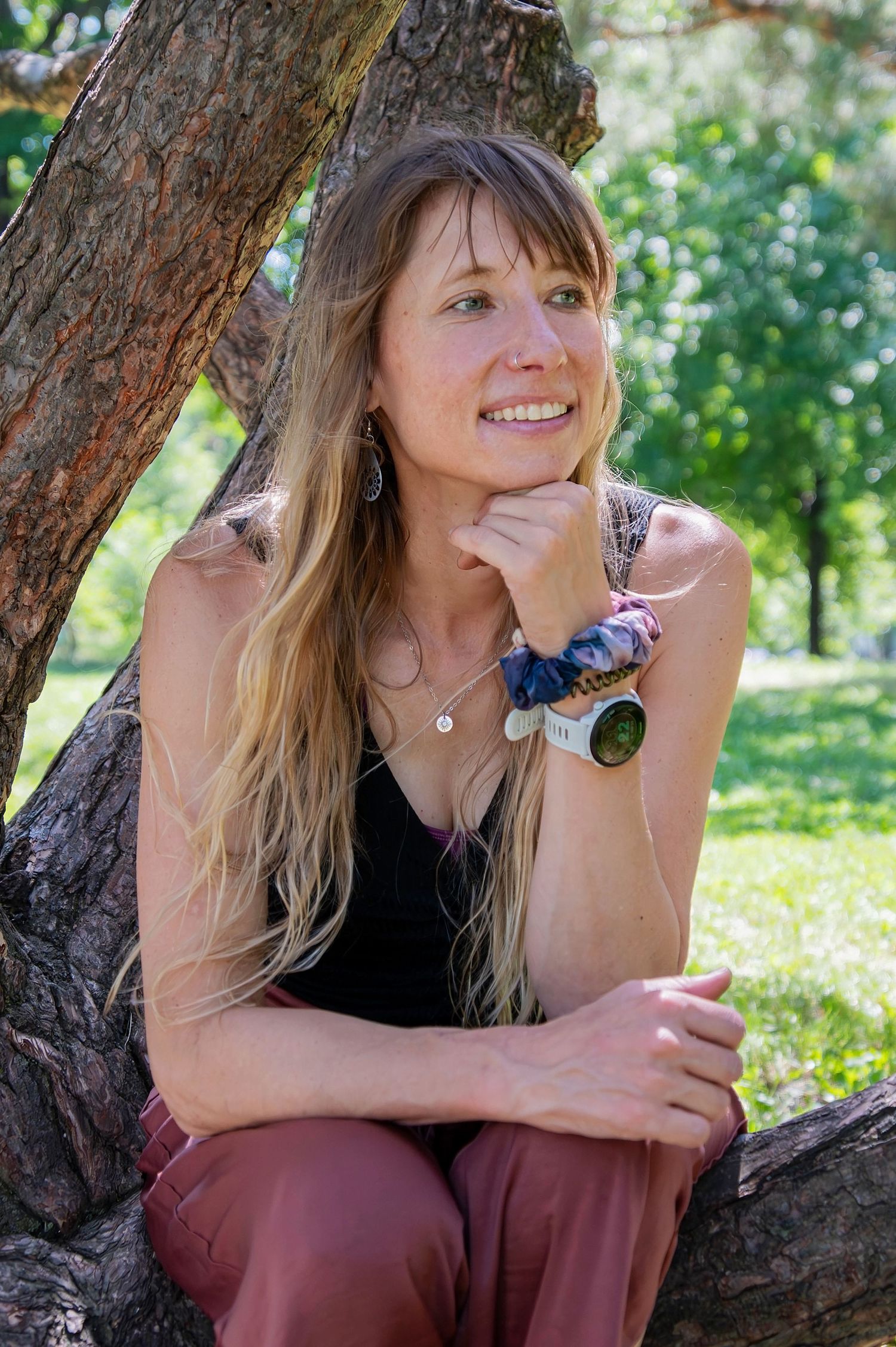 Smiling blonde woman leaning against a tree trunk in a sunny green park setting.