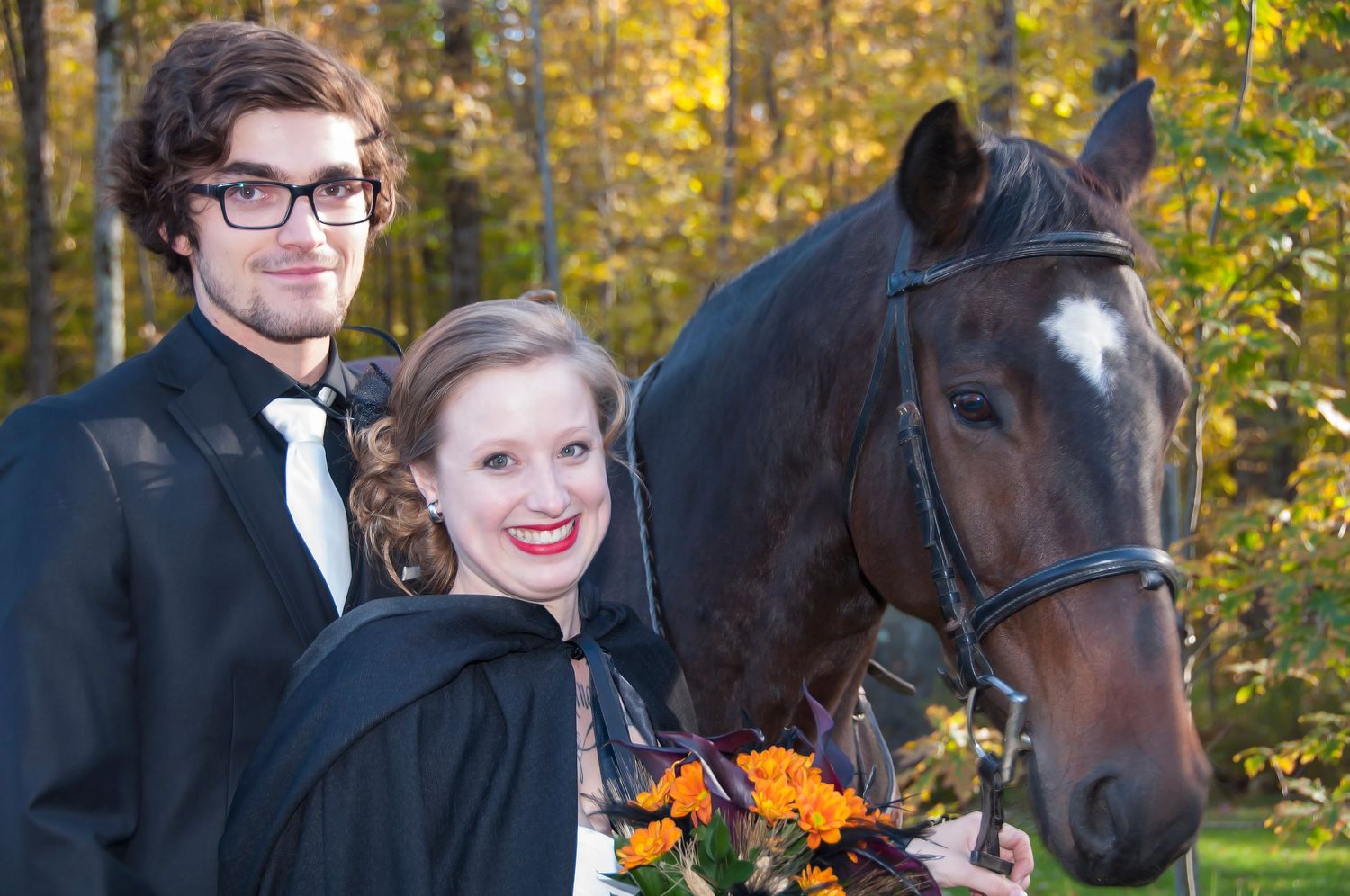 Couple in black graduation gowns smiling with a brown horse amid autumn foliage.