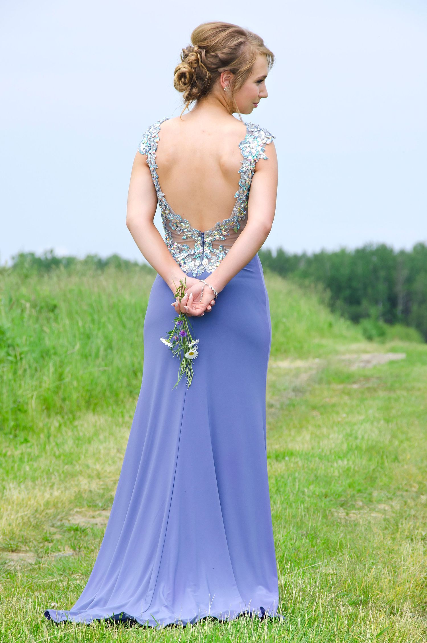 Woman in elegant backless lavender gown holds flowers while standing in green meadow.