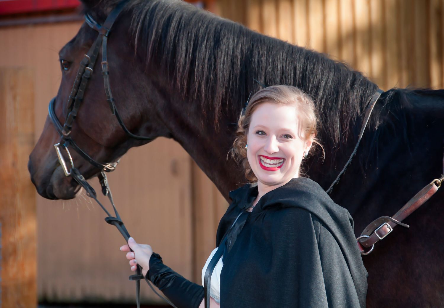 Smiling bride in black cape poses beside a dark horse outside a stable on her wedding day.