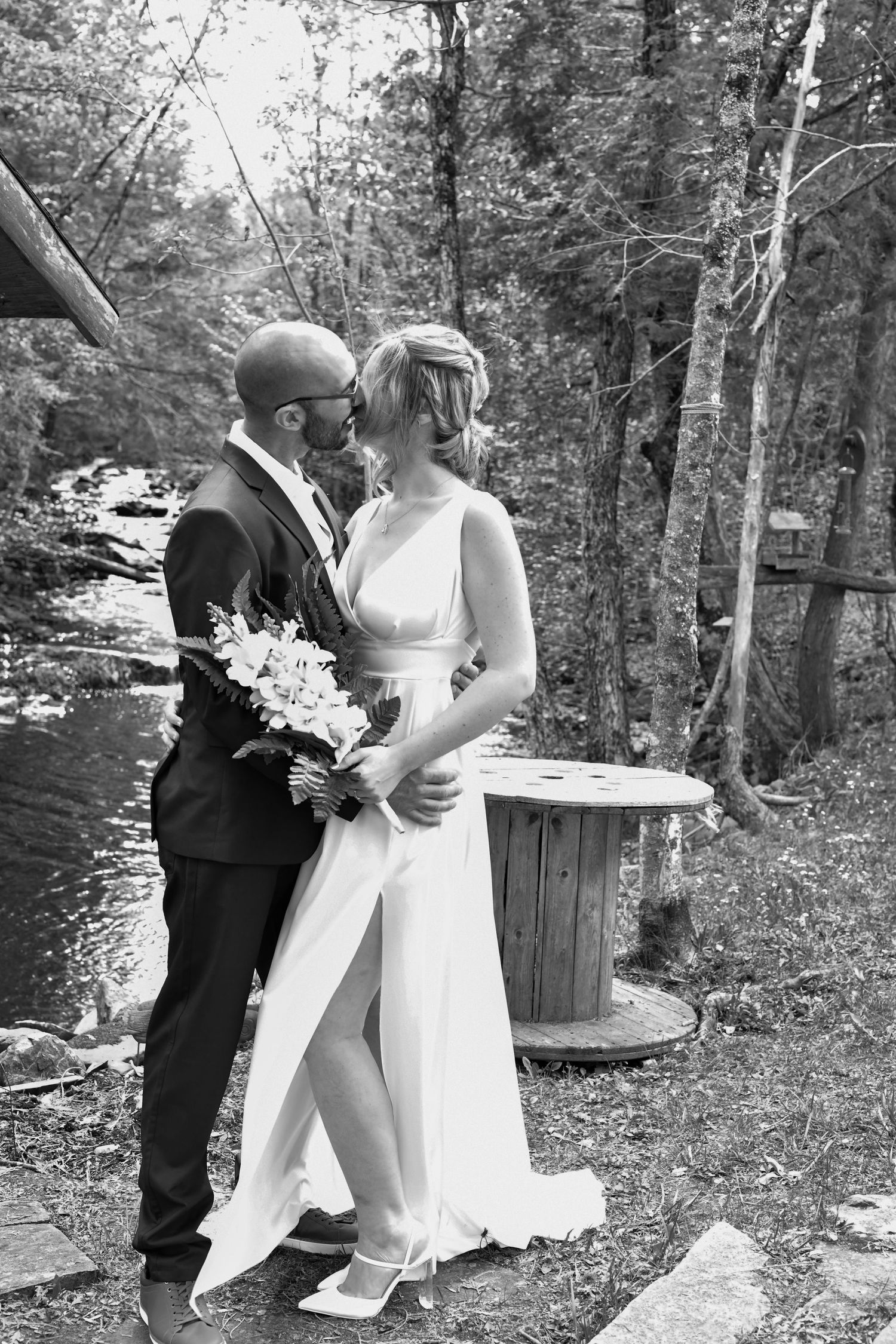 Black and white wedding photo of couple kissing by a forest stream, bride holding a floral bouquet.