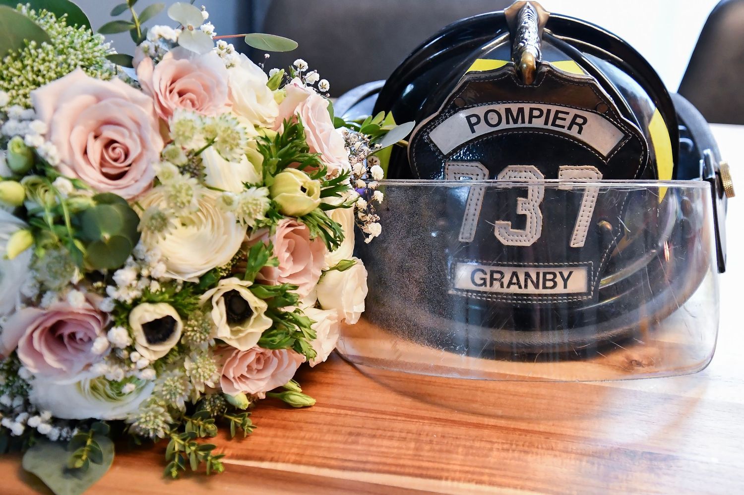 Wedding bouquet of pink and white roses beside a Pompier Granby firefighter helmet numbered 137 on wooden surface.