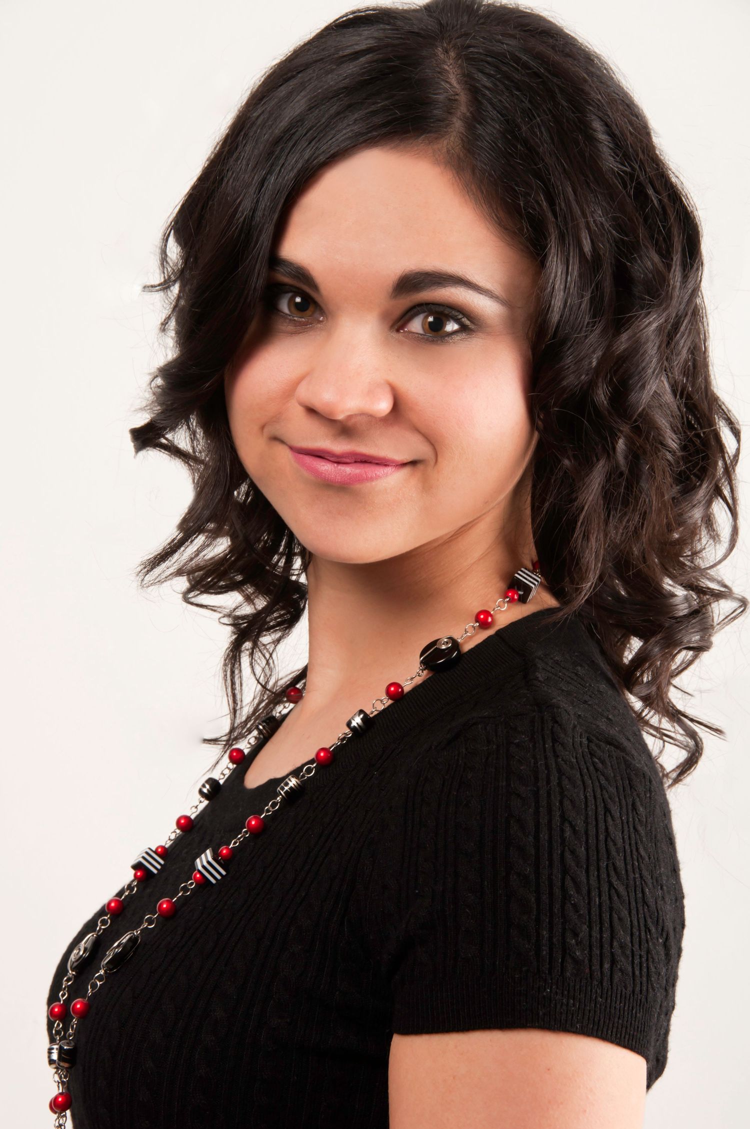 Young woman with dark curly hair wearing black sweater and red beaded necklace smiling at camera.
