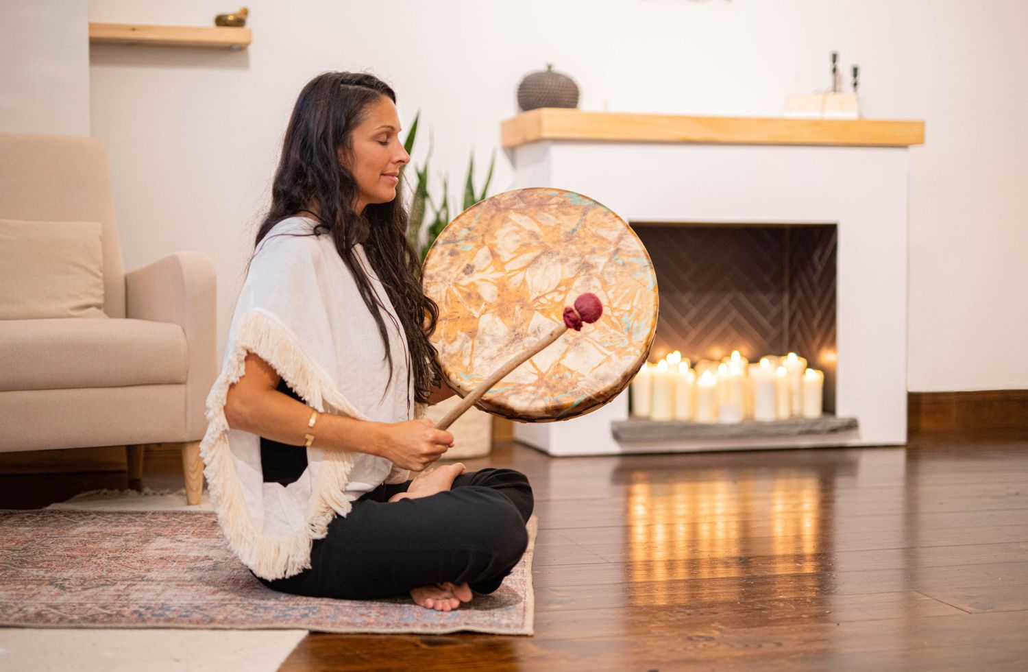 Woman sitting cross-legged playing a shamanic drum near a candlelit fireplace indoors.