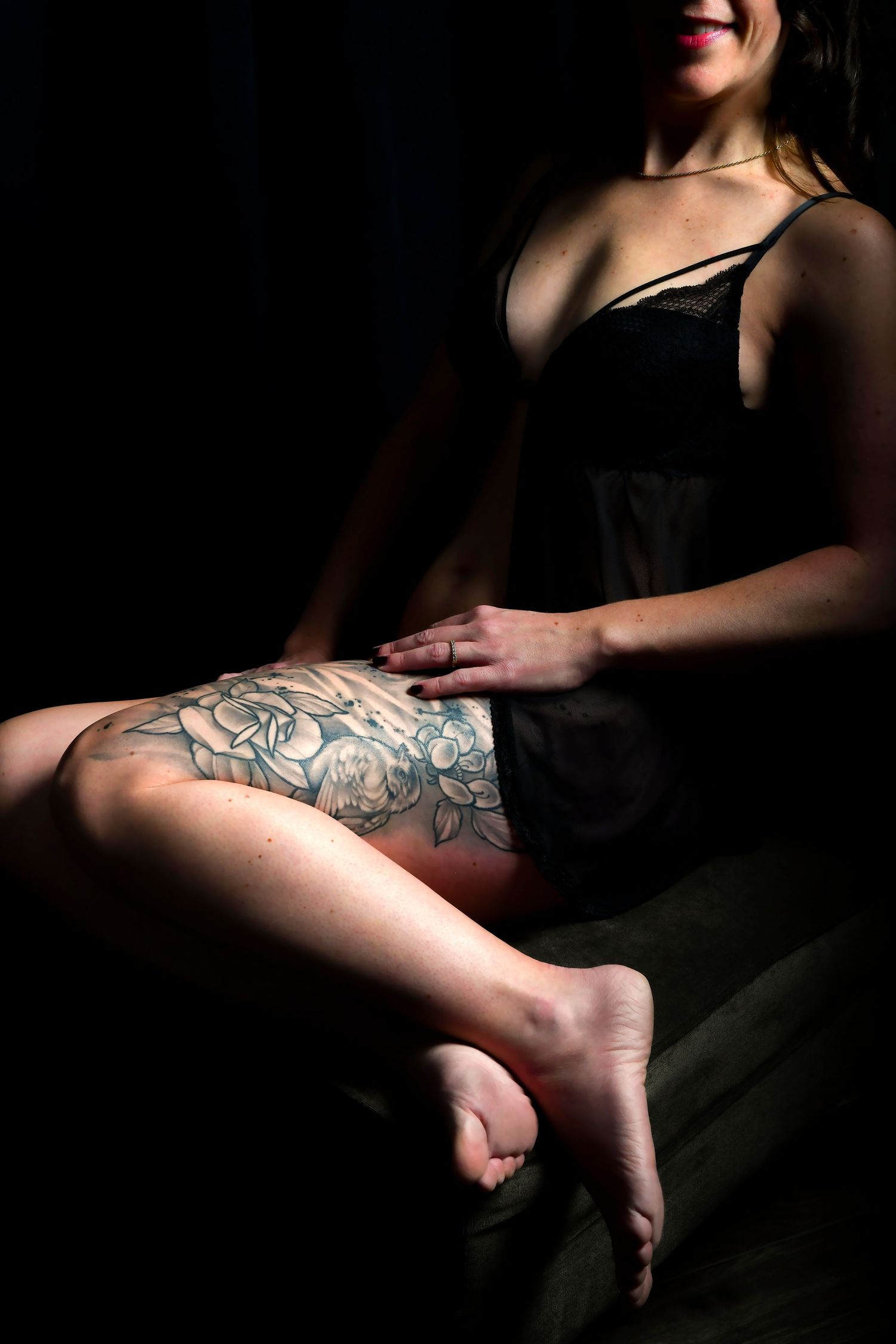 Woman in black lace lingerie seated in shadow showcasing large floral thigh tattoo.