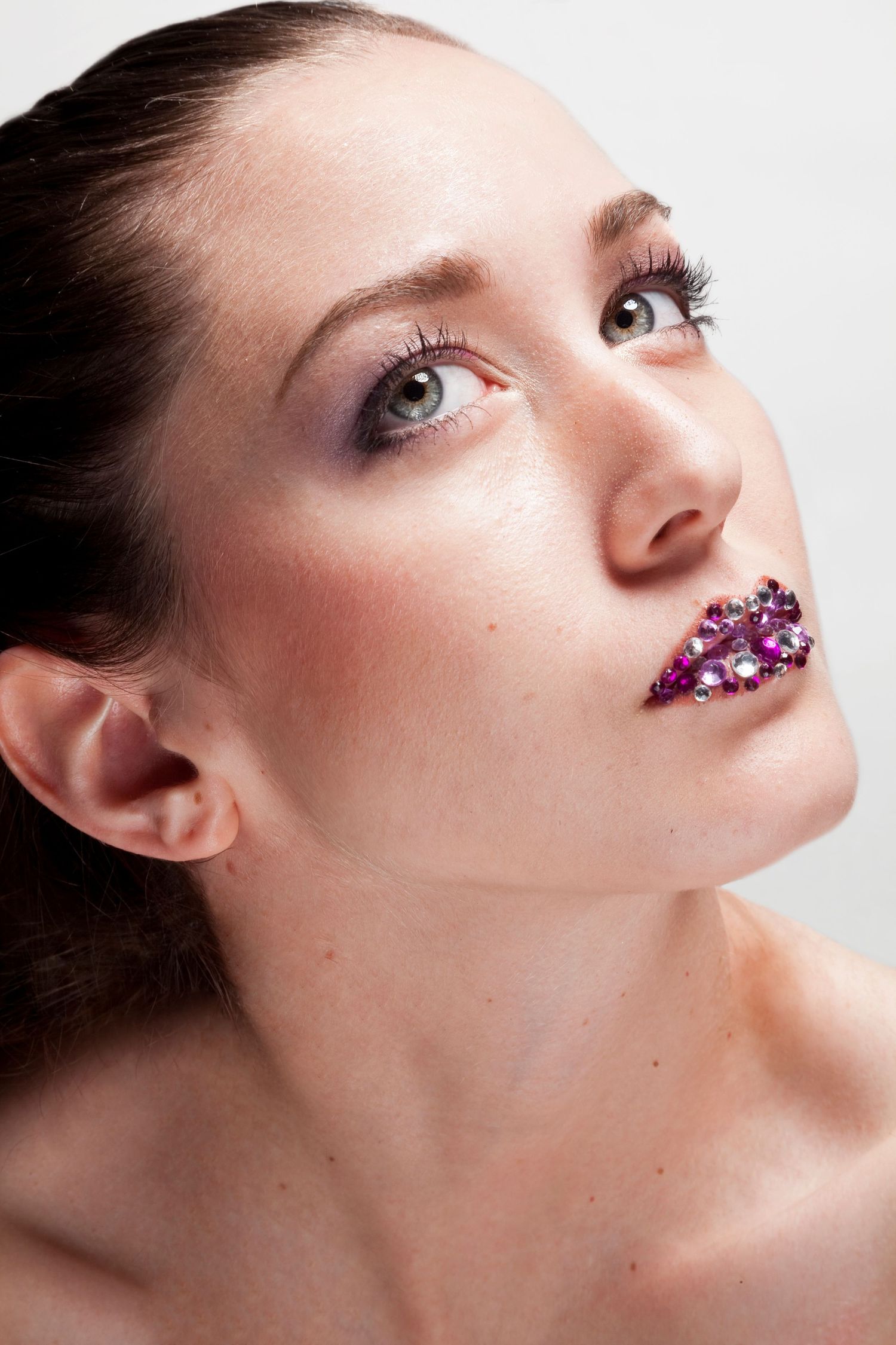 Close-up portrait of woman with pink rhinestone gem lips looking upward against white background.