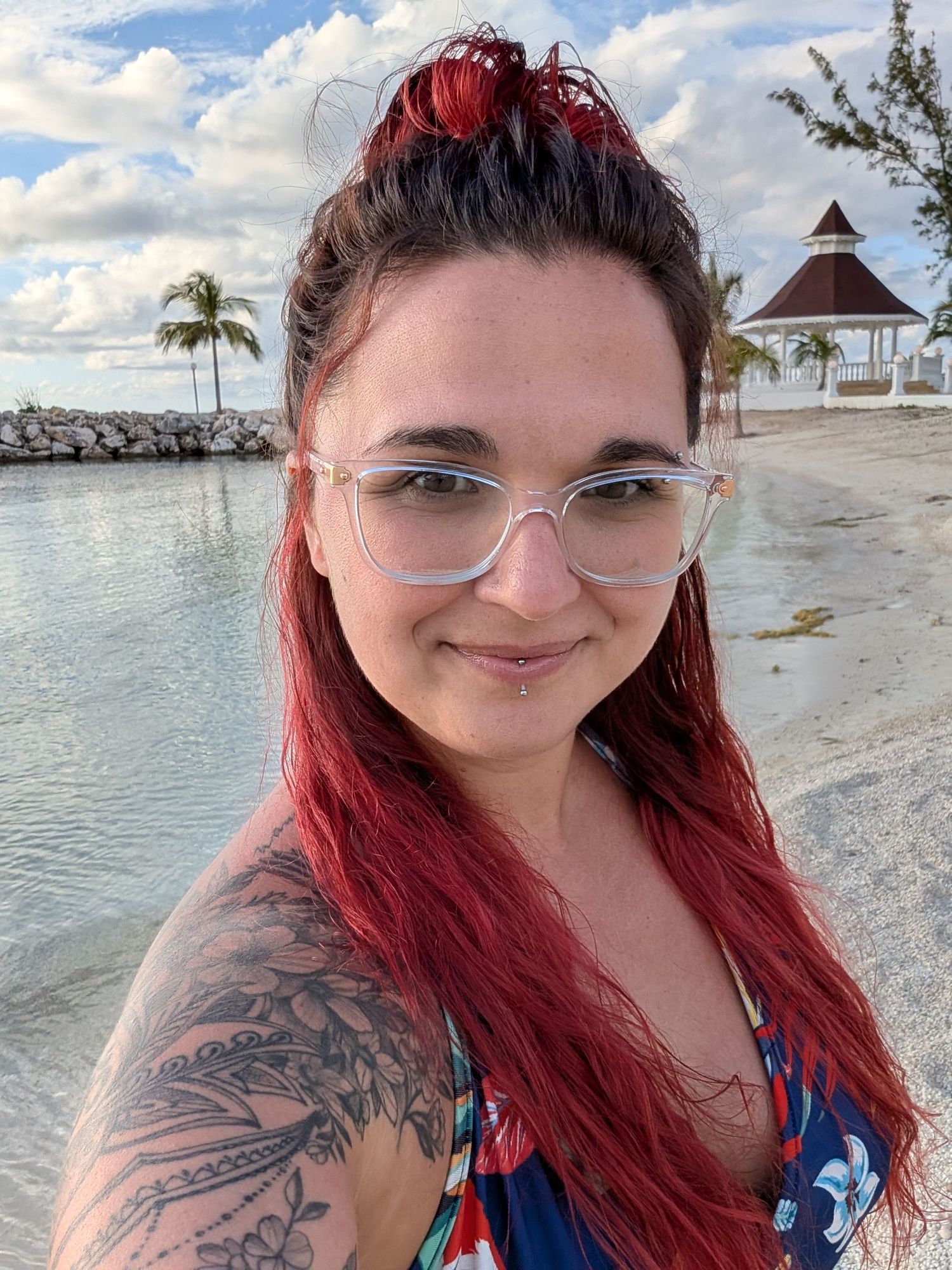 Young woman with red hair and tattoos wearing glasses smiles at beach with gazebo and palm trees.
