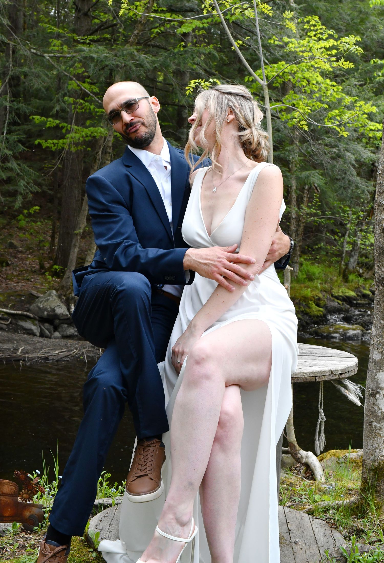 Groom in navy suit and bride in white dress pose together on a dock in a forest setting.