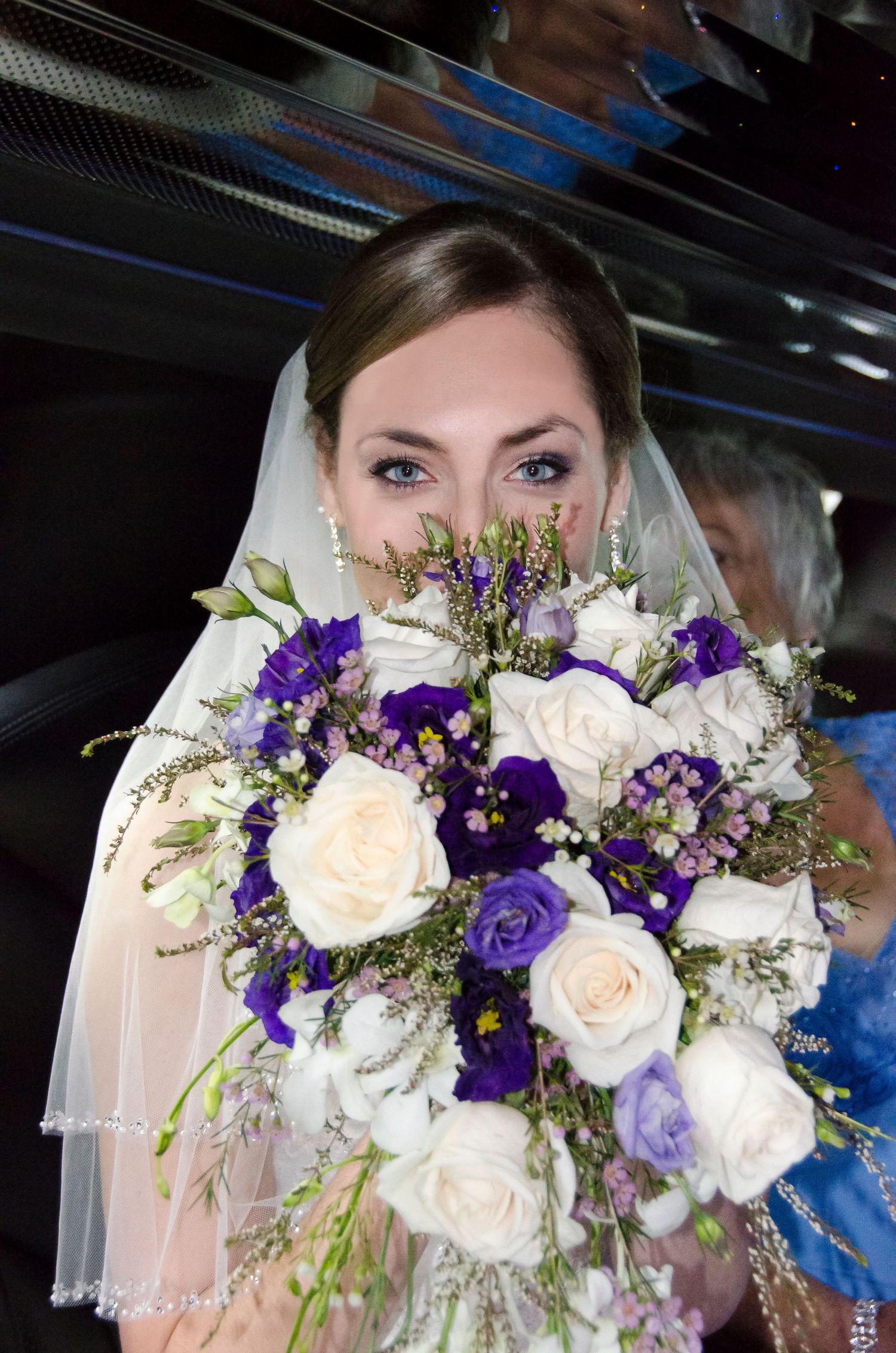 Beautiful bride with blue eyes partially hidden behind a purple and white wedding bouquet.