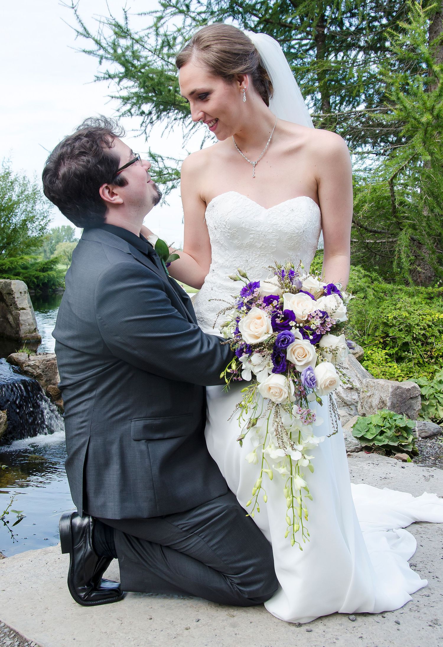 Groom kneels before his tall bride holding a stunning cascading purple and white wedding bouquet.