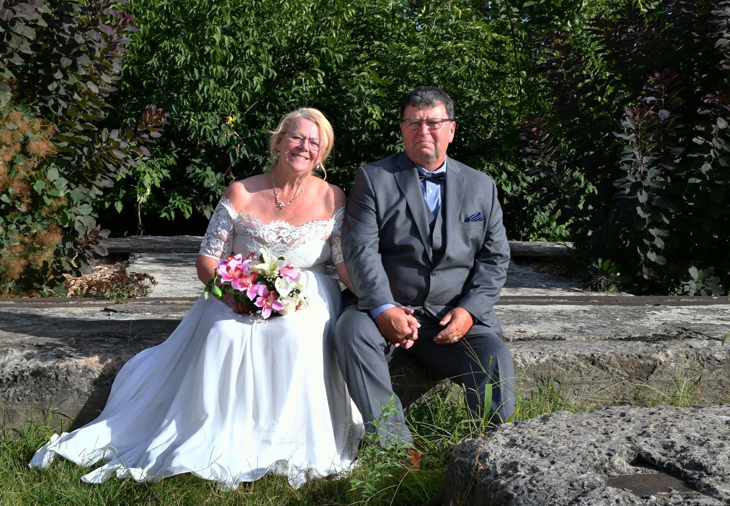 Elegant bride and groom pose together on stone steps surrounded by lush green garden foliage.