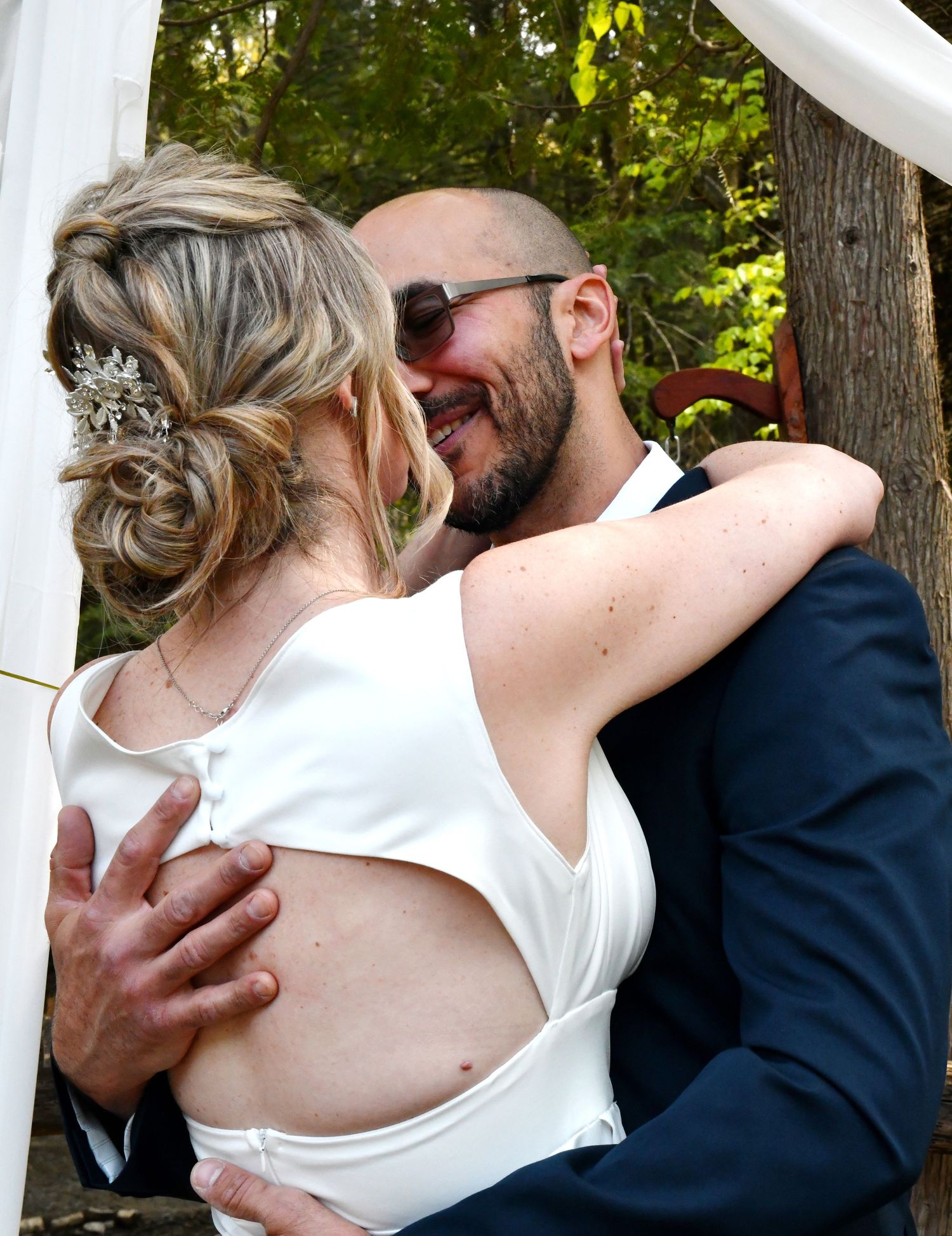 Groom embracing bride from behind during outdoor wedding ceremony under a floral arch in wooded setting.