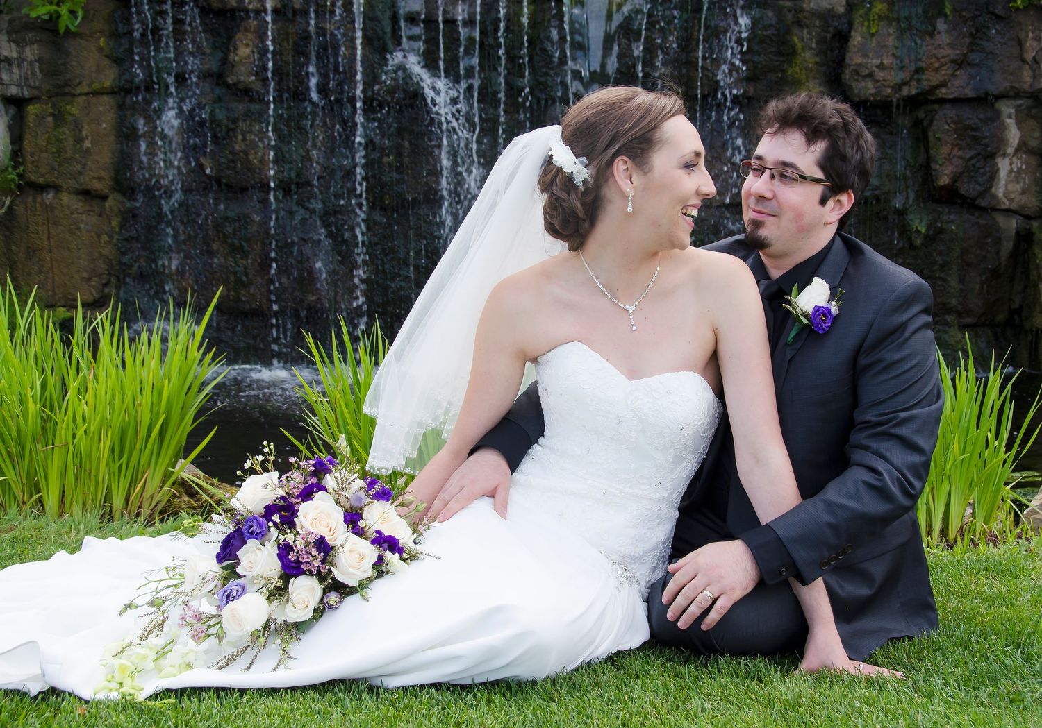 Bride and groom sit together on grass near a waterfall, bride holding purple and white bouquet.