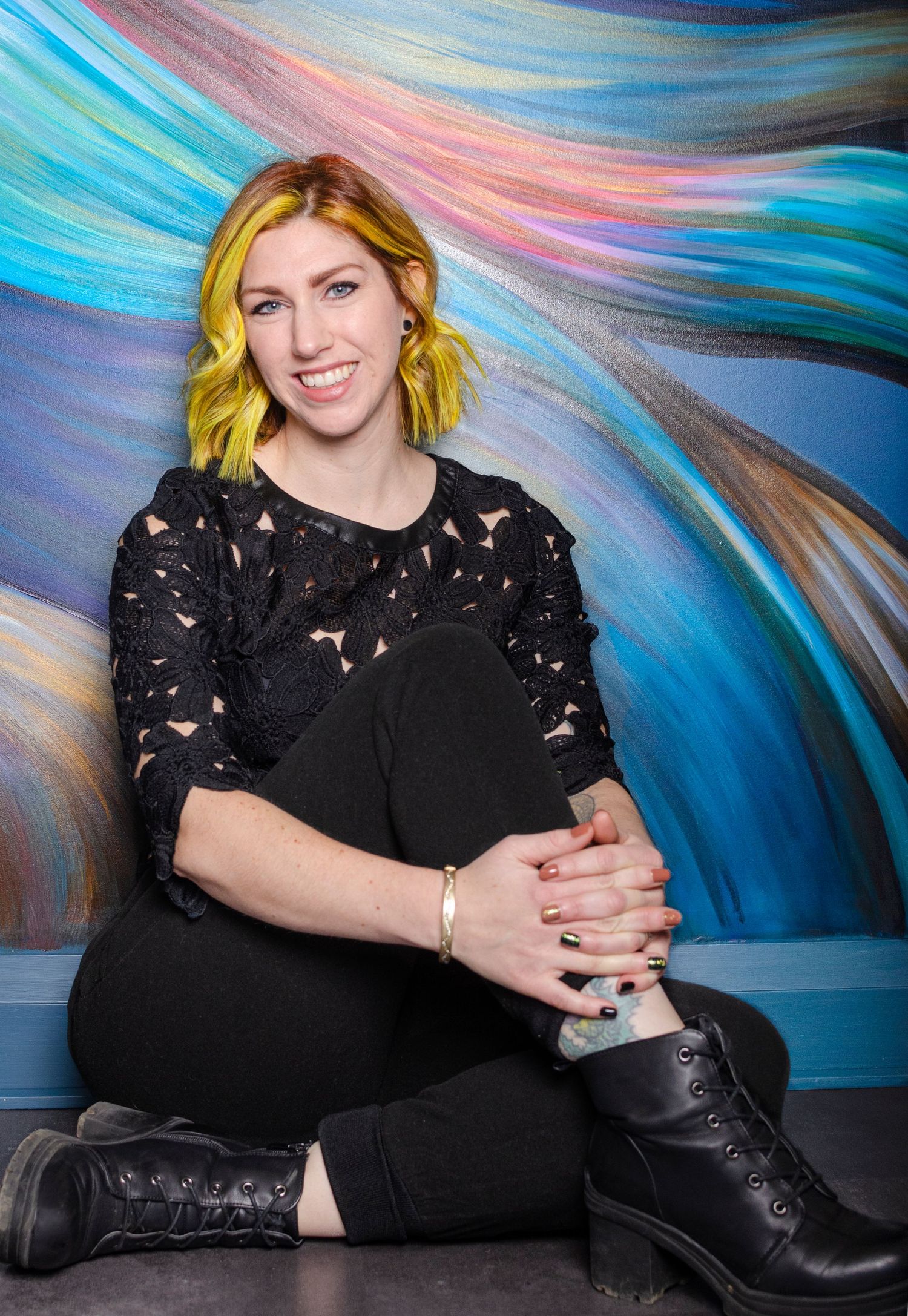 Smiling woman with yellow hair in black outfit sitting against colorful abstract mural background.