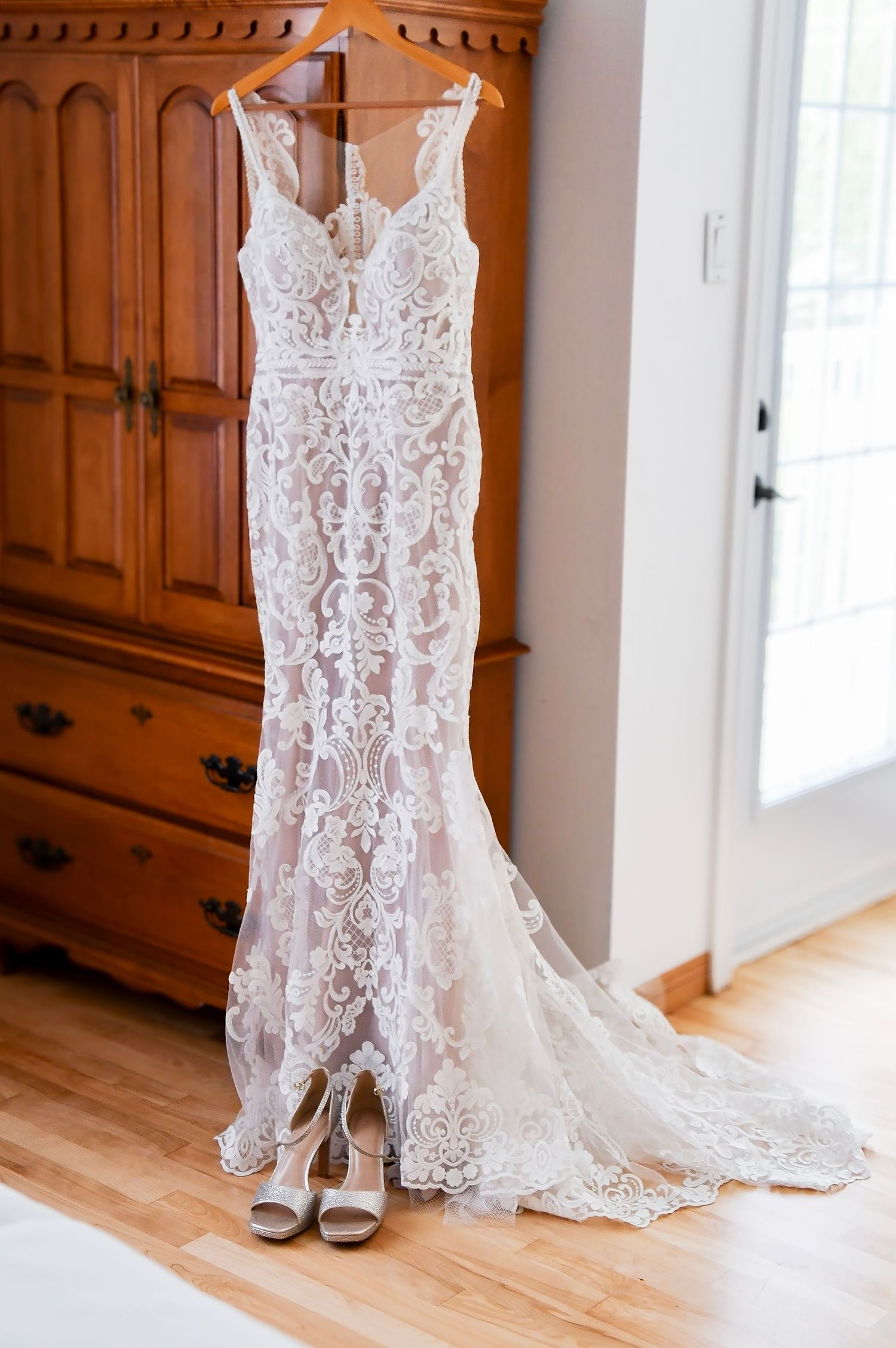 Elegant lace wedding dress hanging on a wooden armoire with bridal heels below.