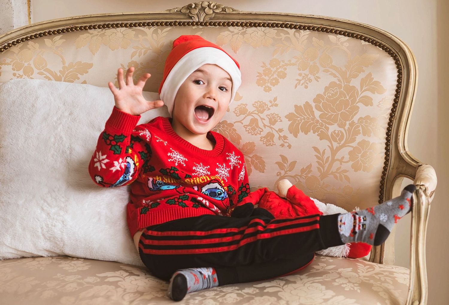 Happy toddler in red Christmas sweater and Santa hat waving while sitting on an ornate sofa.