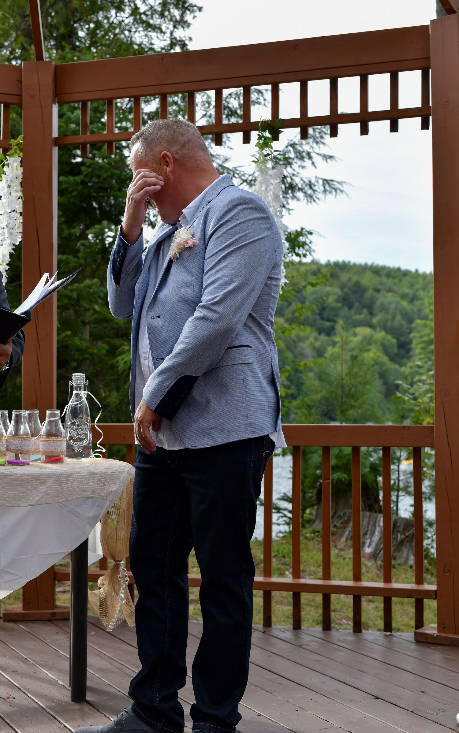 Emotional groom in light blue blazer wiping tears during outdoor wedding ceremony on wooden deck.