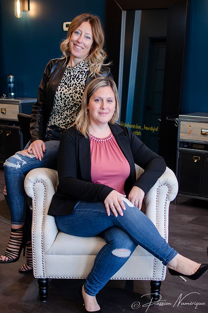 Two smiling women posing in a modern salon, one seated in a white chair and one standing behind her.