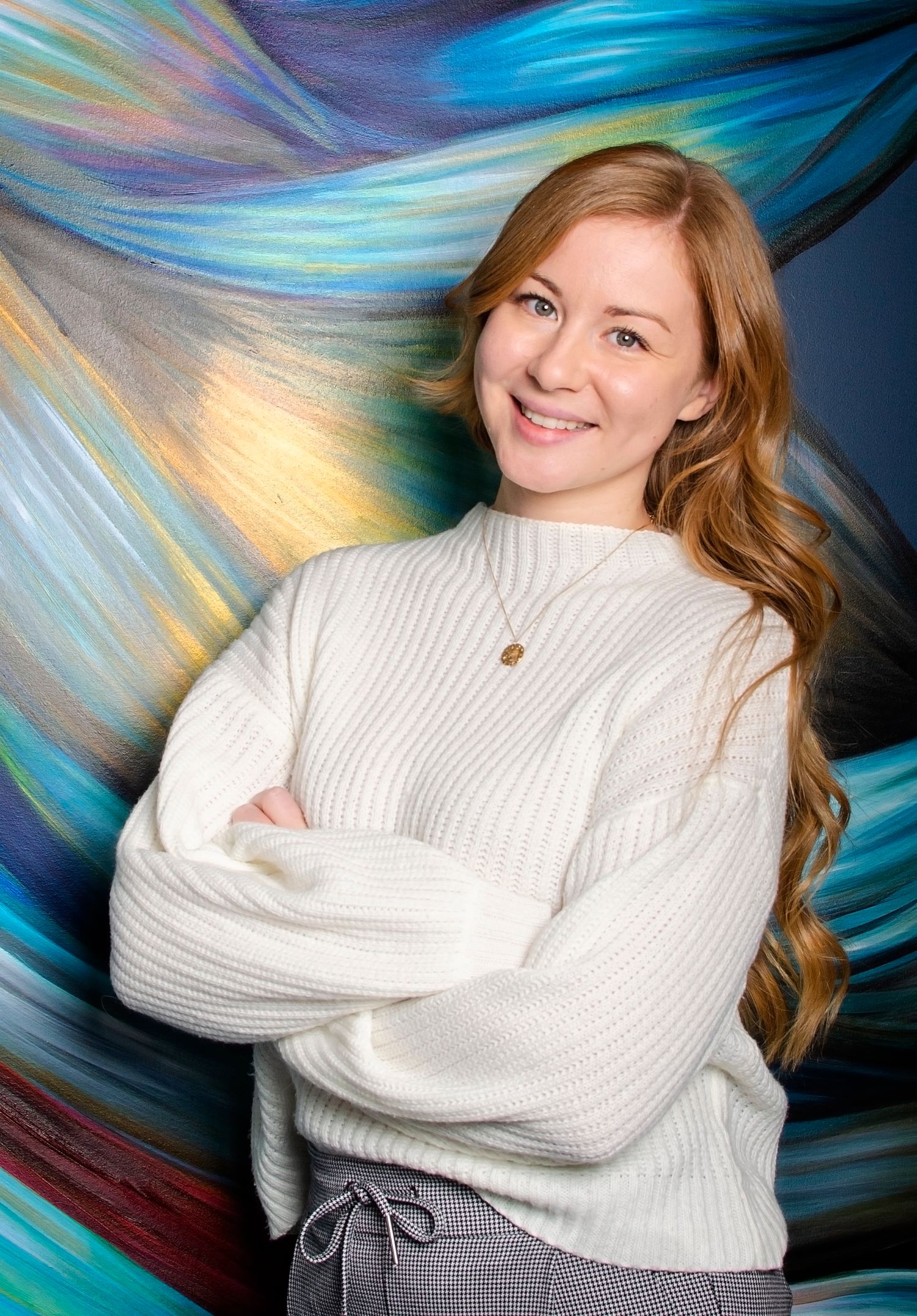Smiling young woman with long red hair posing with arms crossed against a colorful abstract painted background.