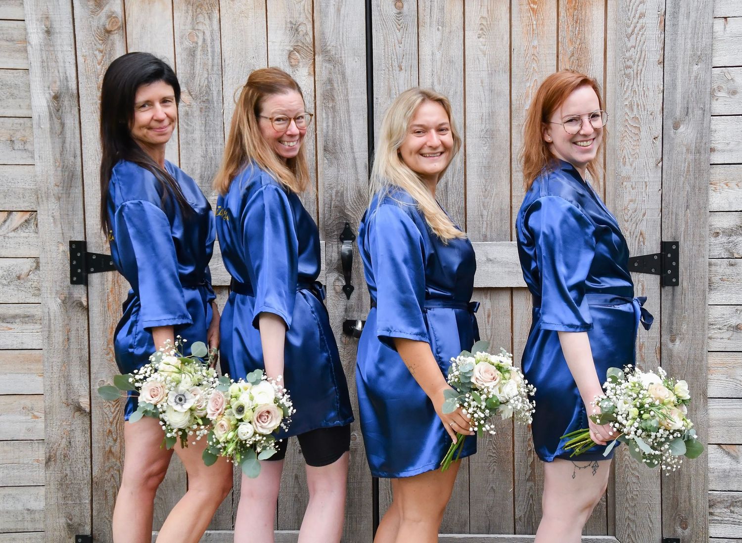 Four bridesmaids in matching navy satin robes holding bouquets in front of a barn door.