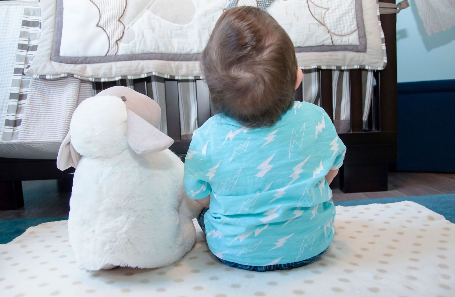 Baby in teal lightning bolt pajamas sitting on floor beside white stuffed lamb toy.