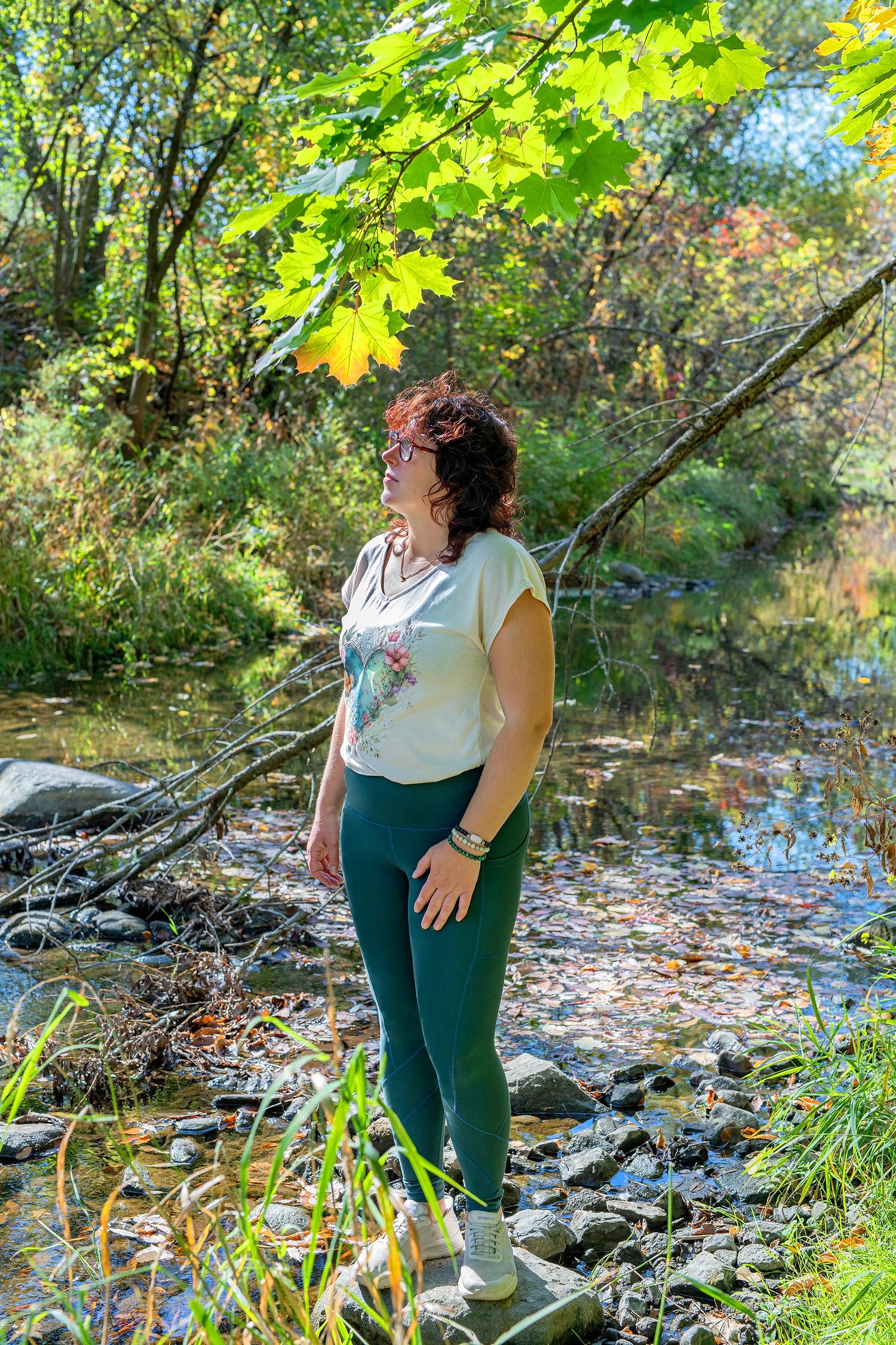 Woman in green leggings and white t-shirt standing beside a rocky stream in an autumn forest.