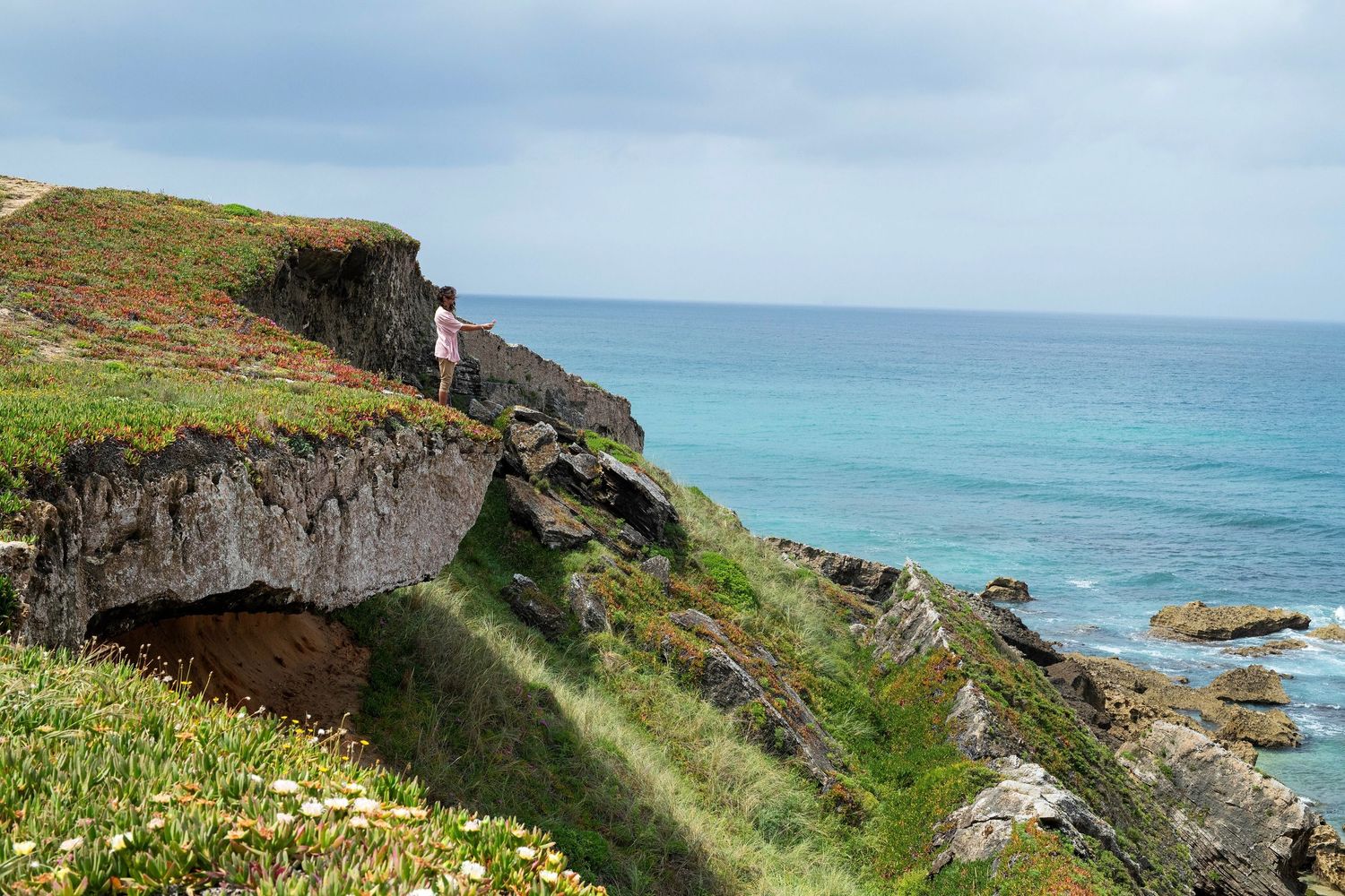 Person standing on dramatic coastal cliffs overlooking a turquoise ocean on a cloudy day.