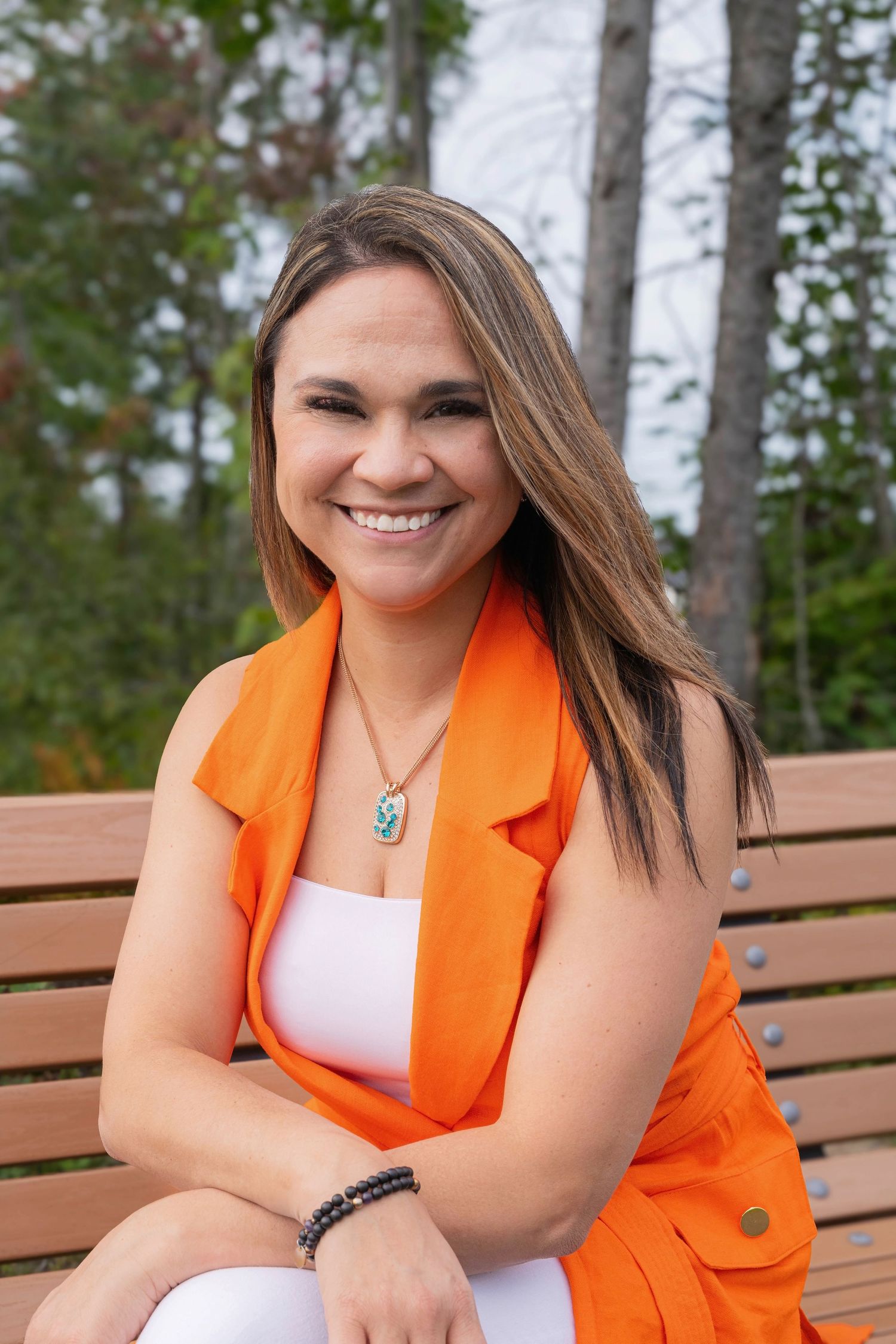 Smiling woman in orange vest sits on wooden bench outdoors among tall trees.