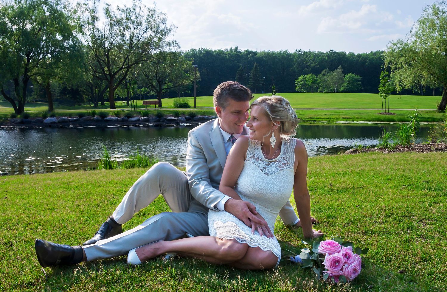Bride and groom sitting on lush green grass by a pond, sharing a tender moment on their wedding day.
