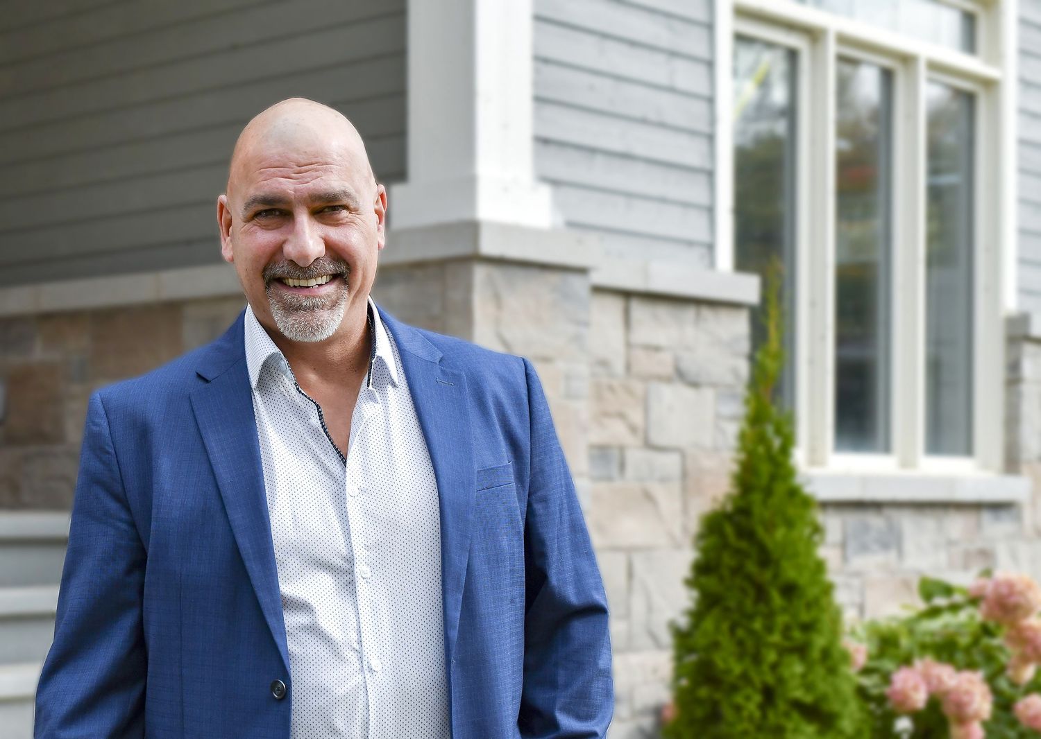 Smiling bald man in blue blazer standing outside modern home with stone and gray siding.