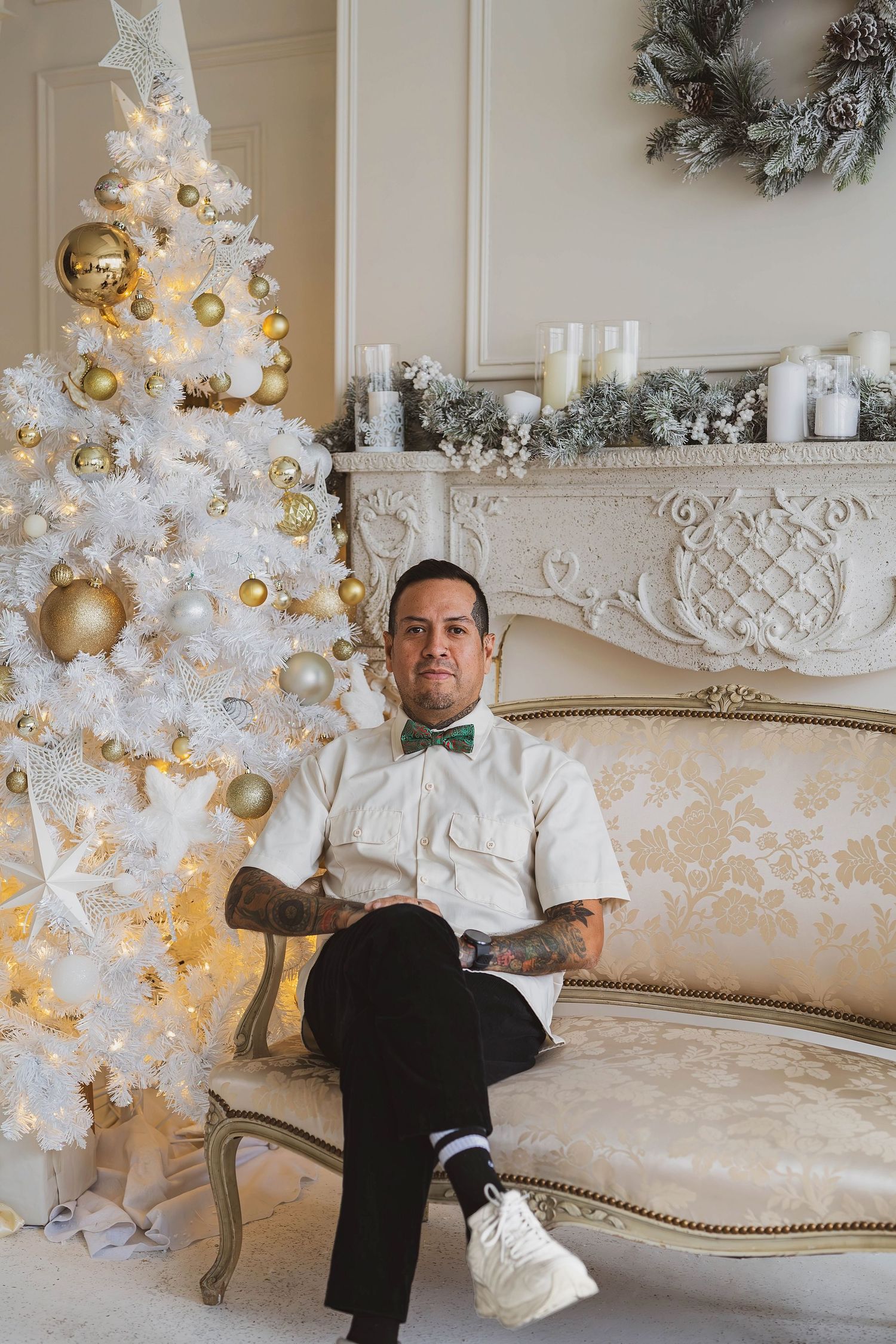 Tattooed man in white shirt and green bow tie sitting on elegant sofa near decorated white Christmas tree.