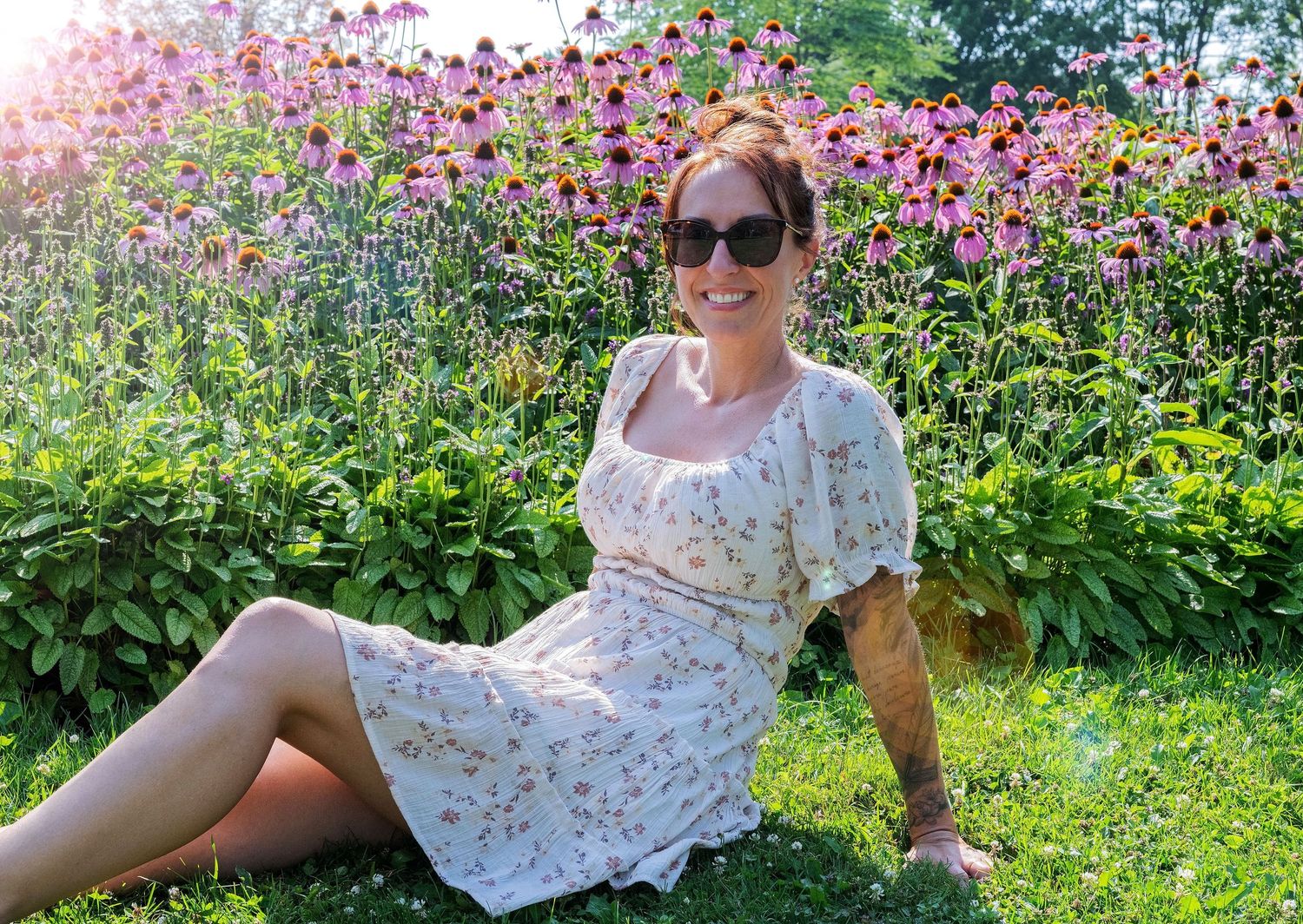 Smiling woman in floral dress sitting by pink echinacea flowers in summer garden.