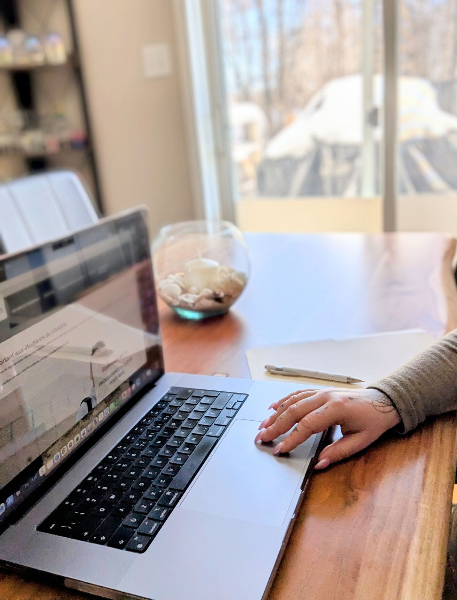 Person working on laptop at wooden desk near bright window with notepad and decorative bowl nearby.