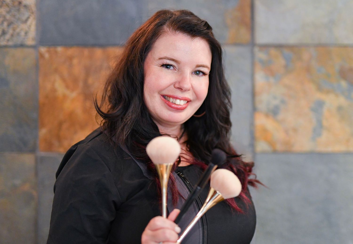 Smiling female makeup artist holding large powder brushes against a stone tile background.
