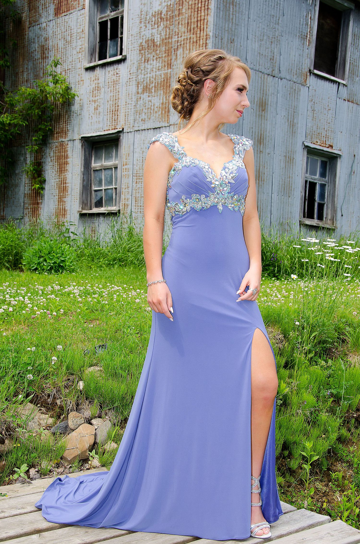 Young woman in lavender formal gown with lace bodice poses near rustic building outdoors.