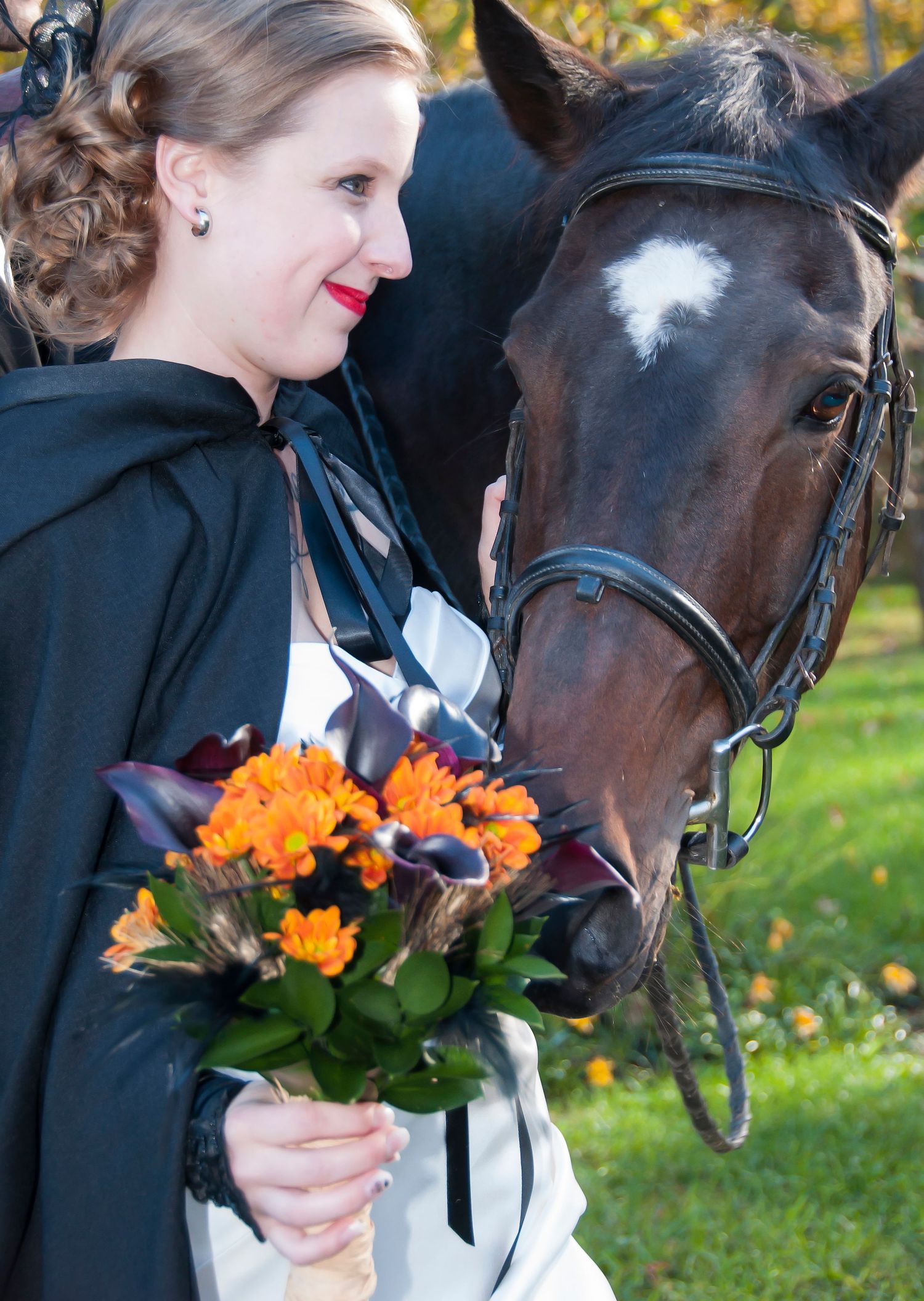 Elegant bride in black cape holds vibrant orange bouquet while standing beside a dark horse outdoors.