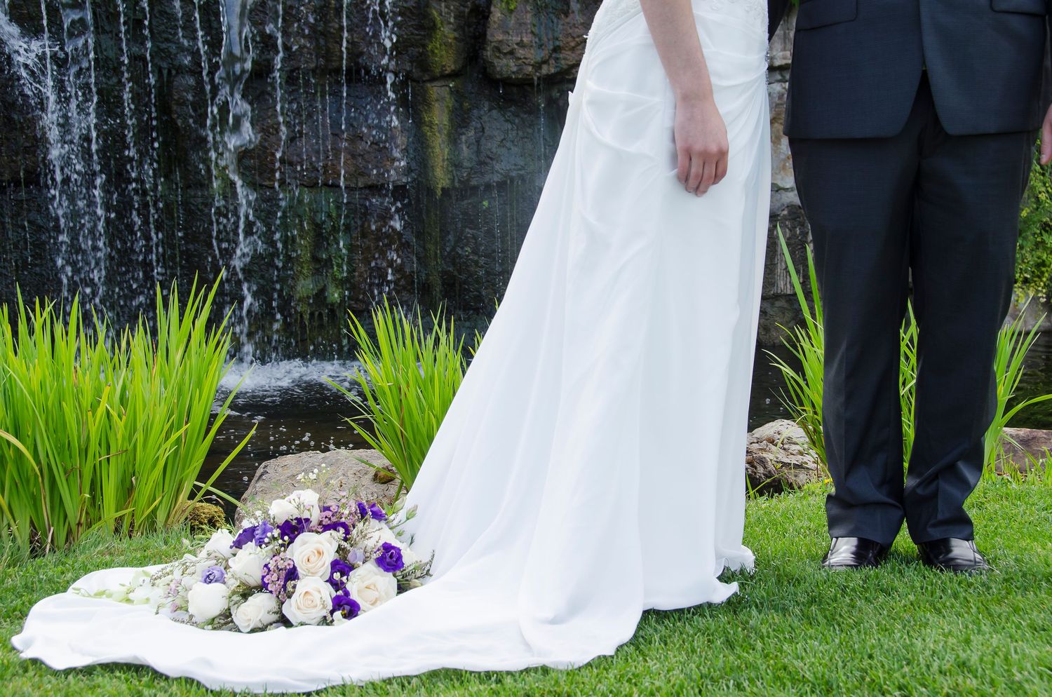 Close-up of a bride's white wedding gown train with purple and white bouquet on green grass.