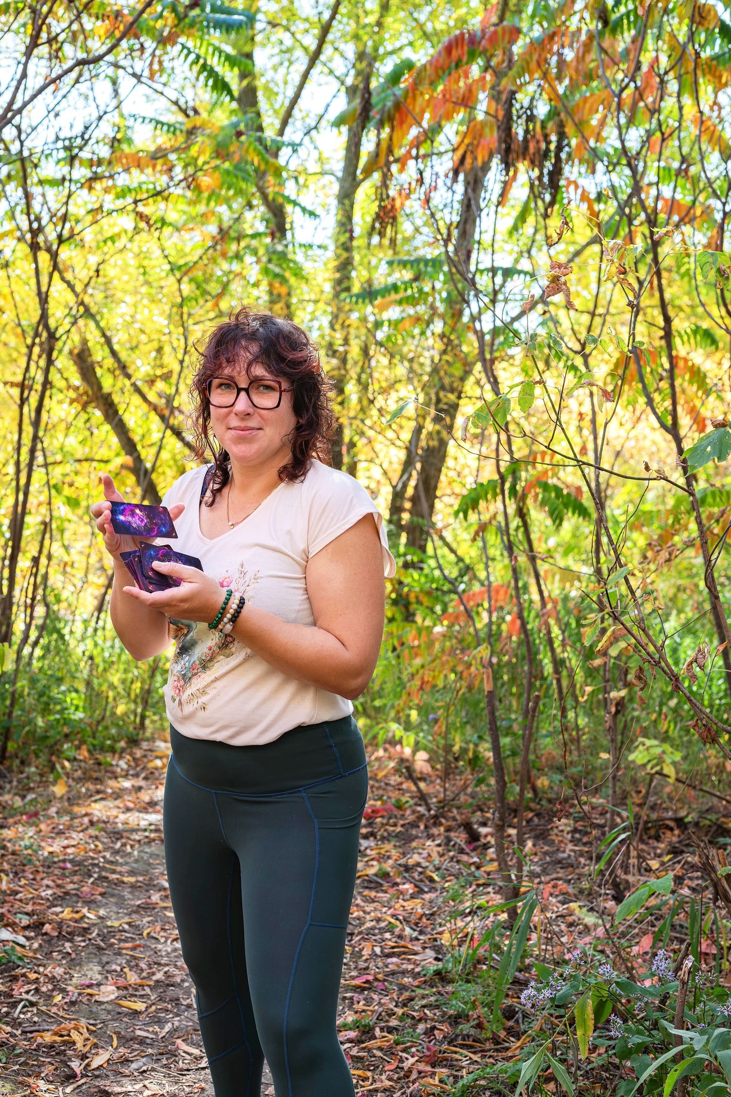 Woman with glasses reading book on autumn forest trail surrounded by colorful foliage.