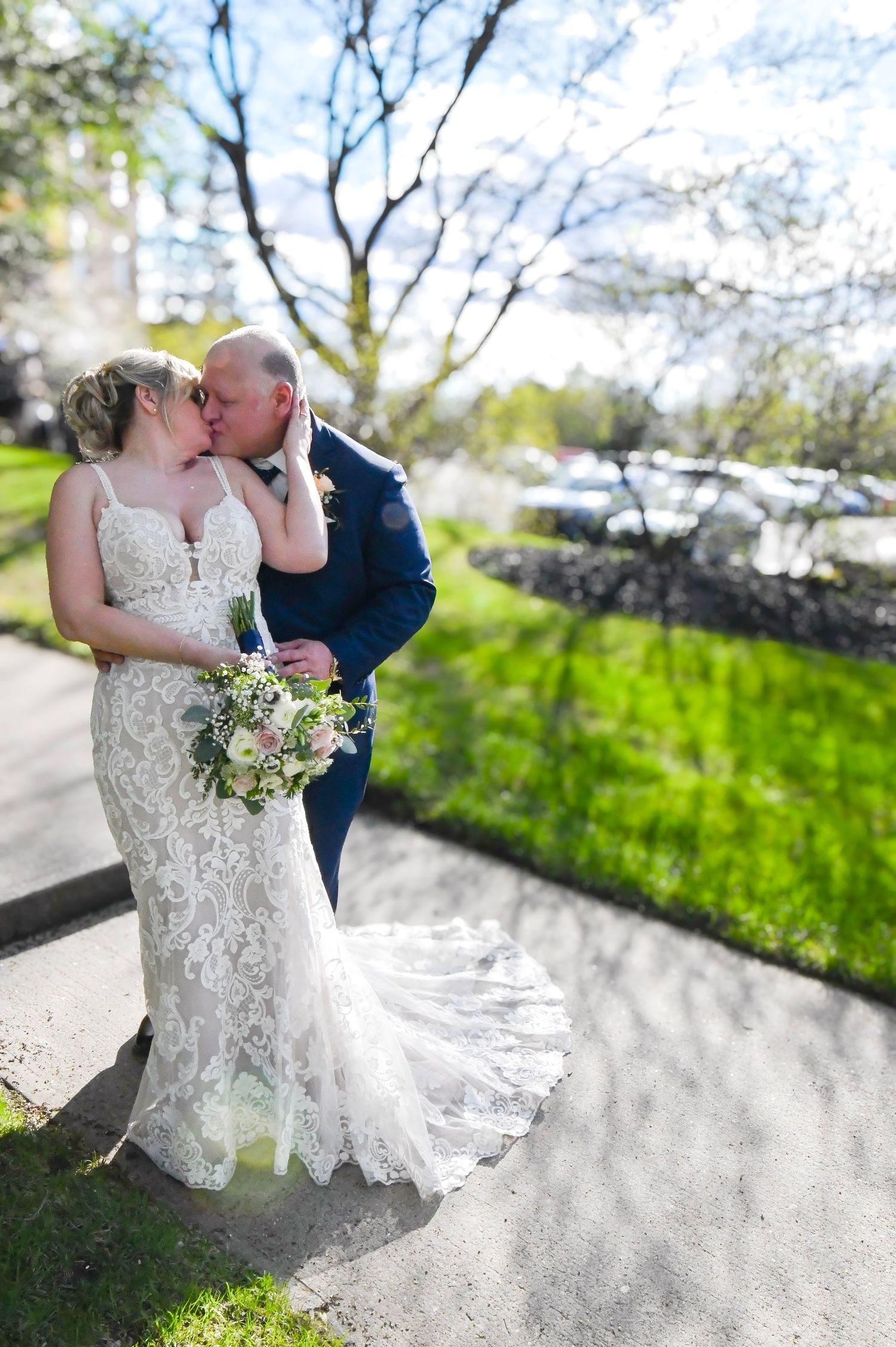 Bride and groom sharing a kiss outdoors in spring, bride holding a floral bouquet in a lace wedding gown.