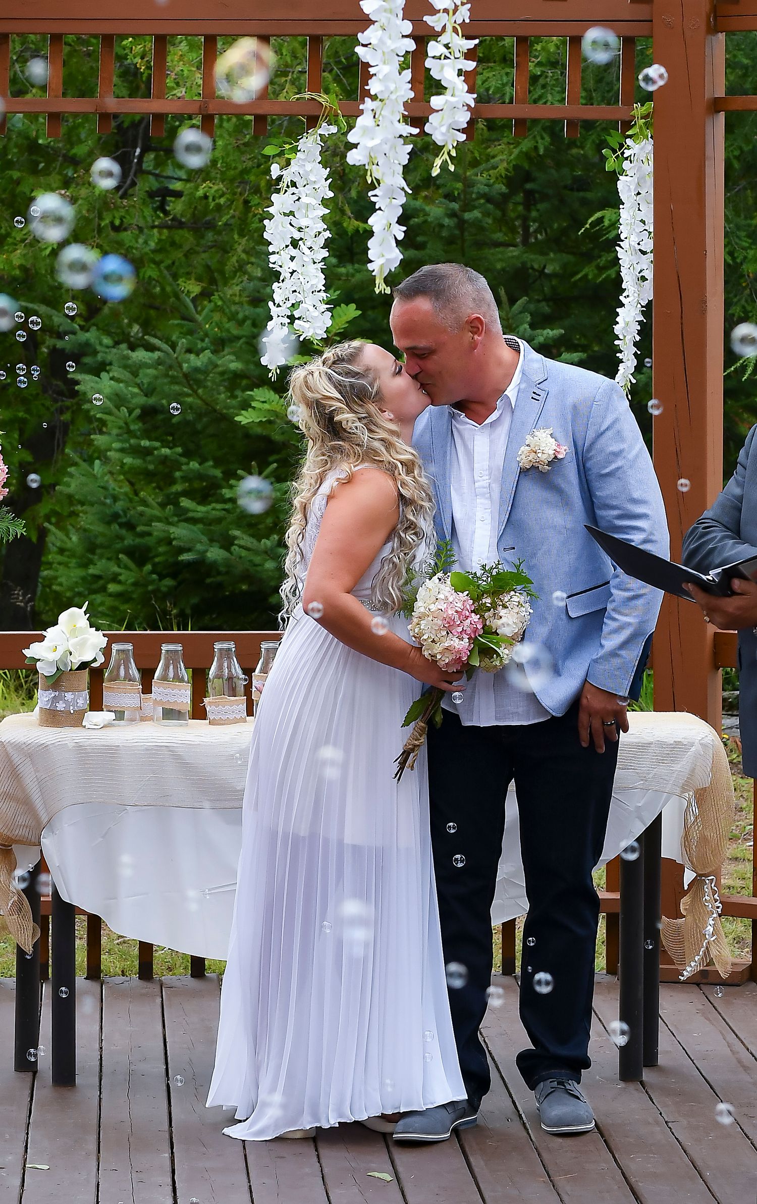 Bride and groom share first kiss under floral arch surrounded by bubbles at outdoor wedding ceremony.