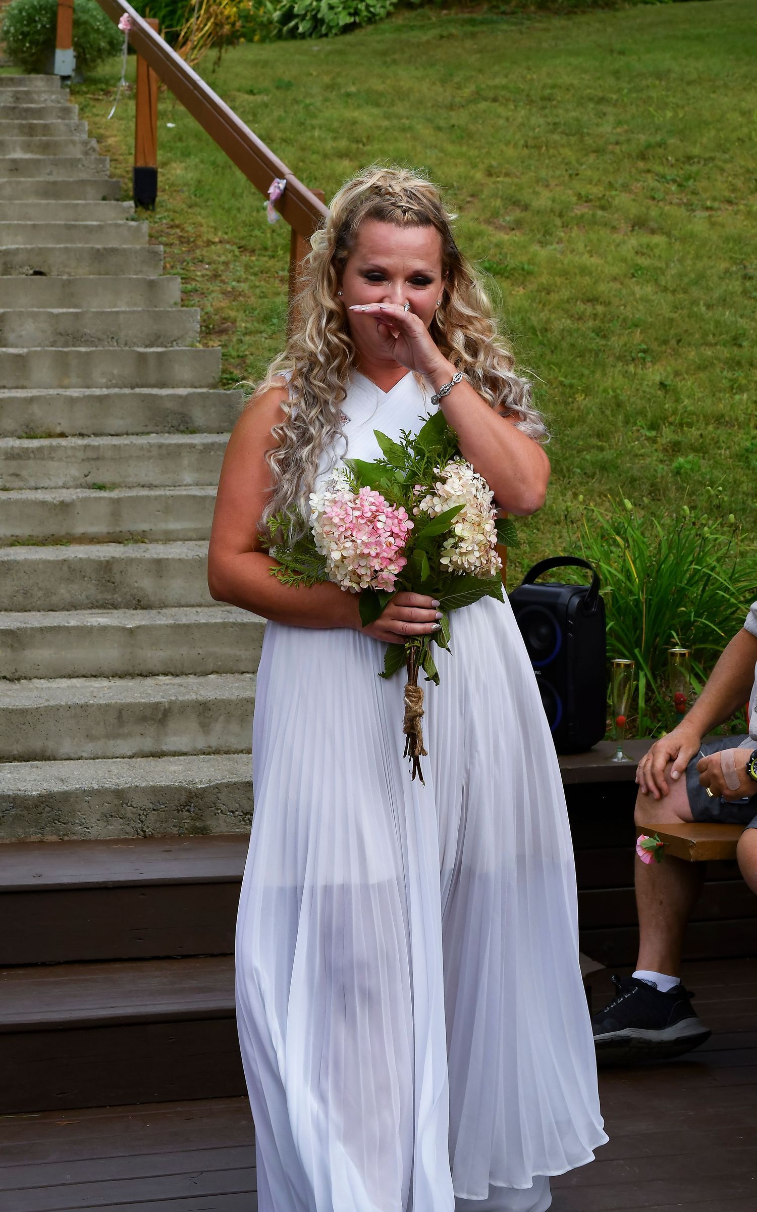 Emotional bride in white pleated dress holding pink and white hydrangea bouquet on outdoor staircase.