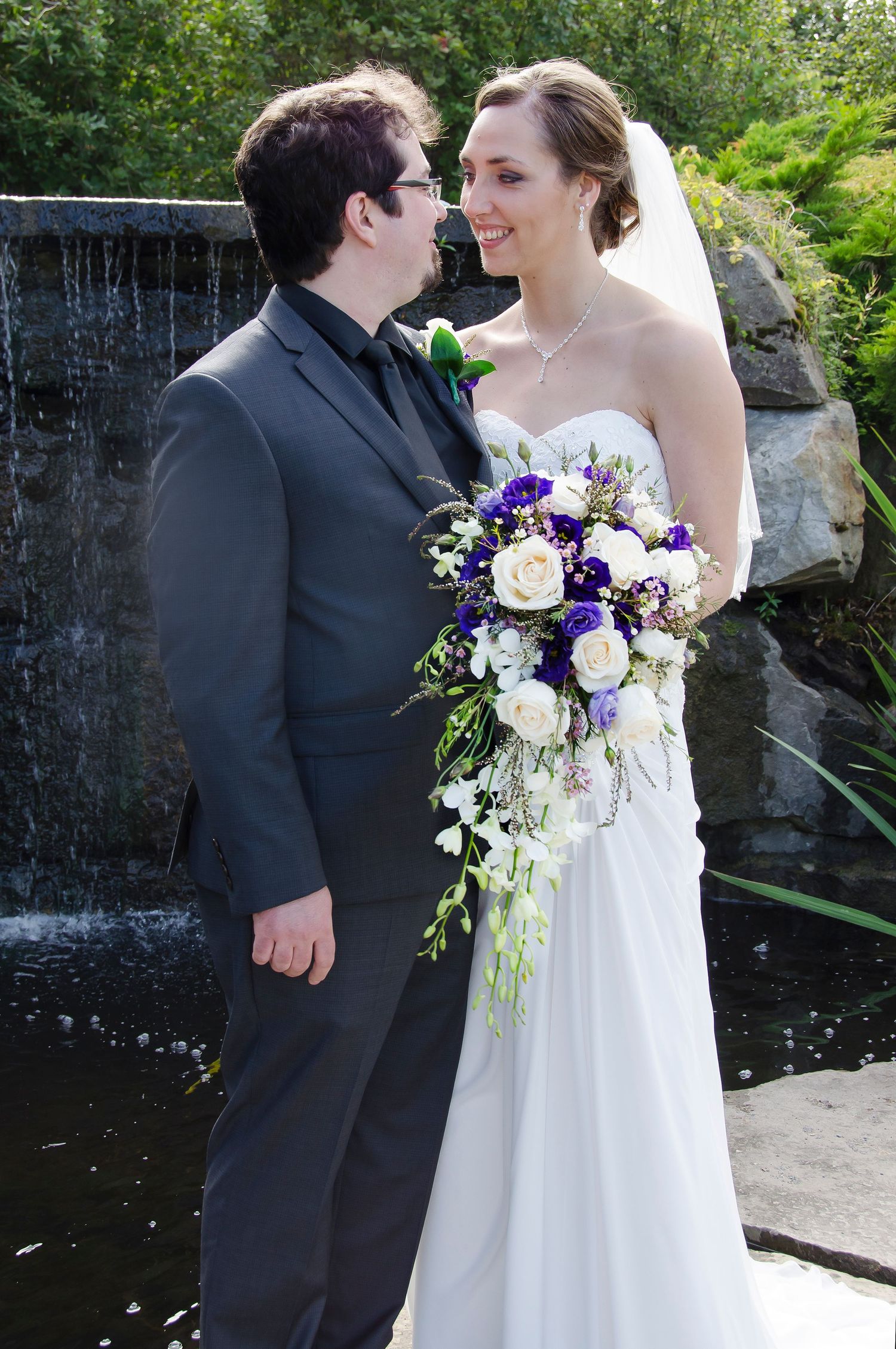 Smiling bride and groom stand together near a waterfall, bride holding a cascading floral bouquet.