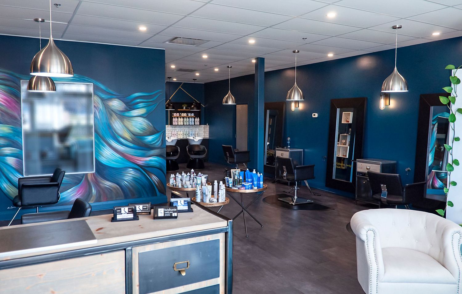 Modern hair salon interior with teal walls, pendant lights, styling chairs, and a vibrant abstract mural.