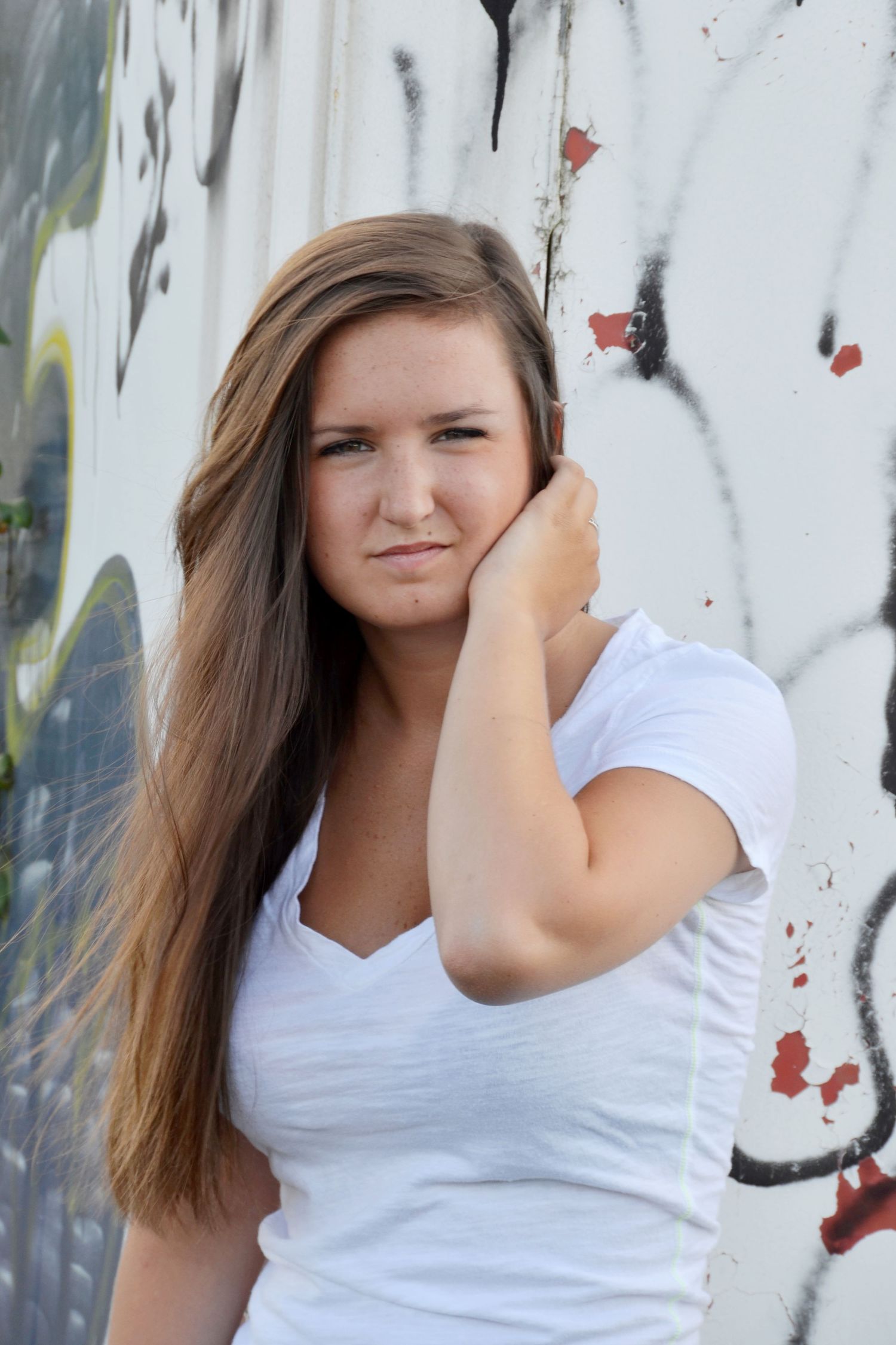 Young woman with long brown hair poses against colorful graffiti wall in white t-shirt.
