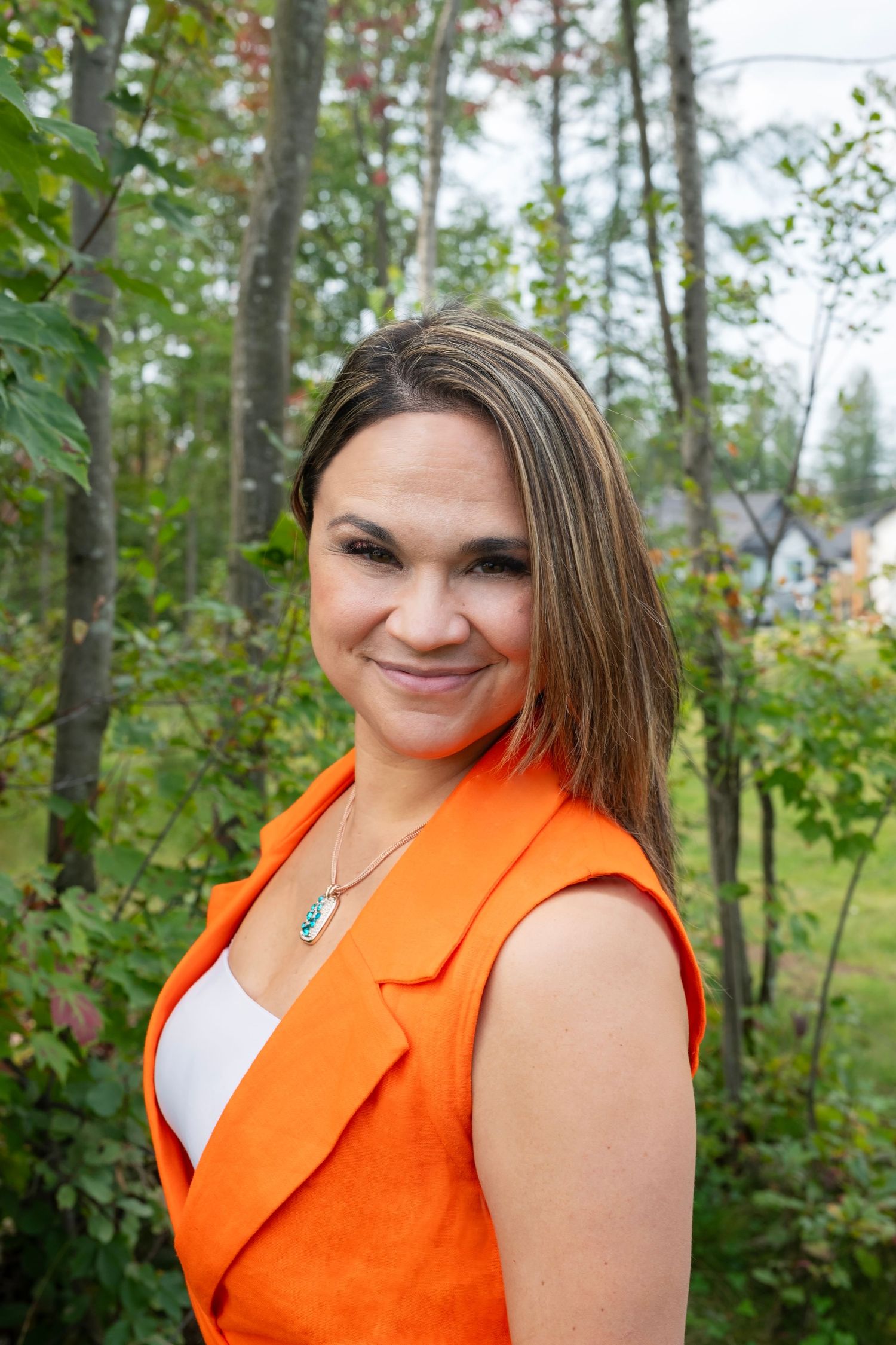 Smiling woman in an orange vest standing outdoors among lush green trees in summer.