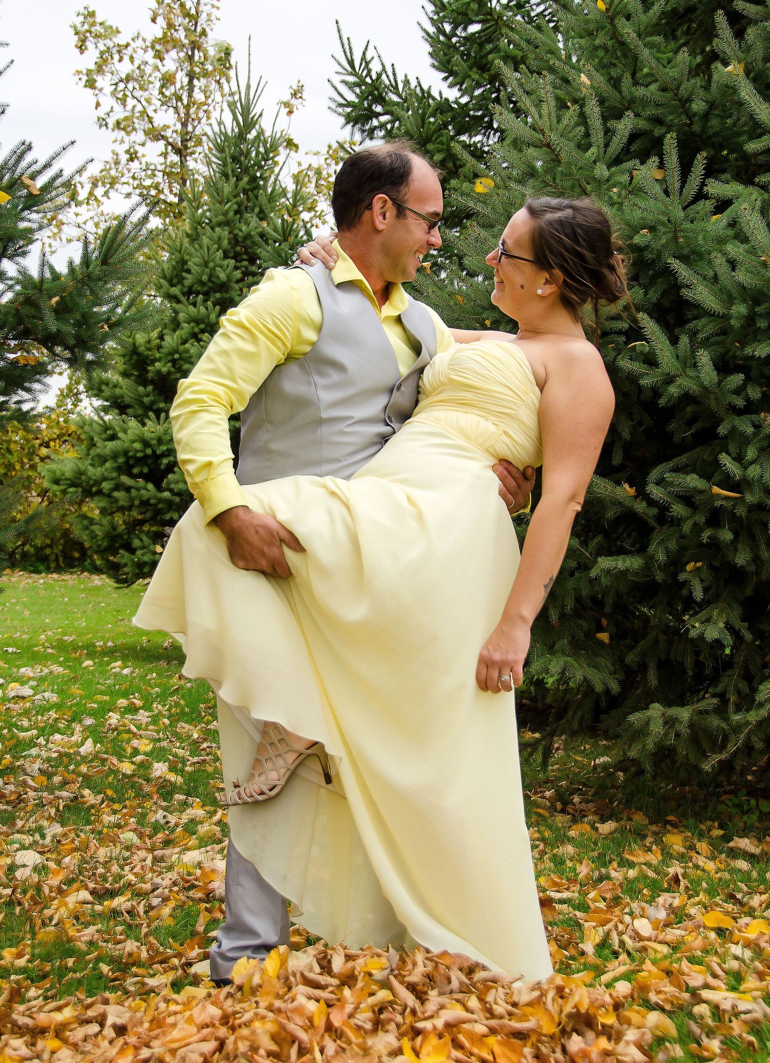 Groom lifting bride in yellow dress among autumn leaves with evergreen trees in the background.