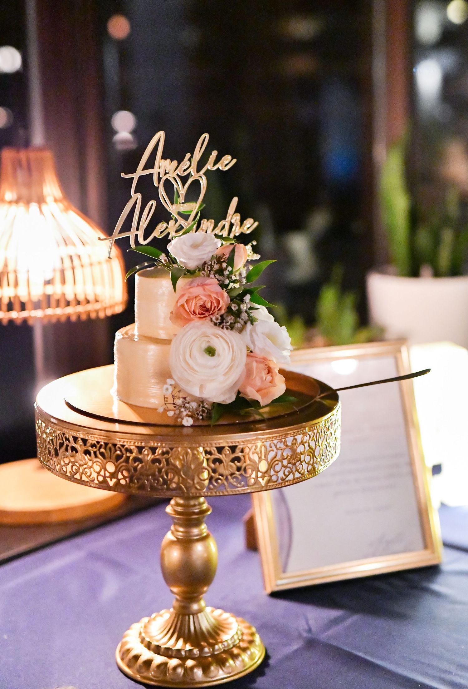 Two-tier wedding cake with floral decor and custom name topper on a gold cake stand.
